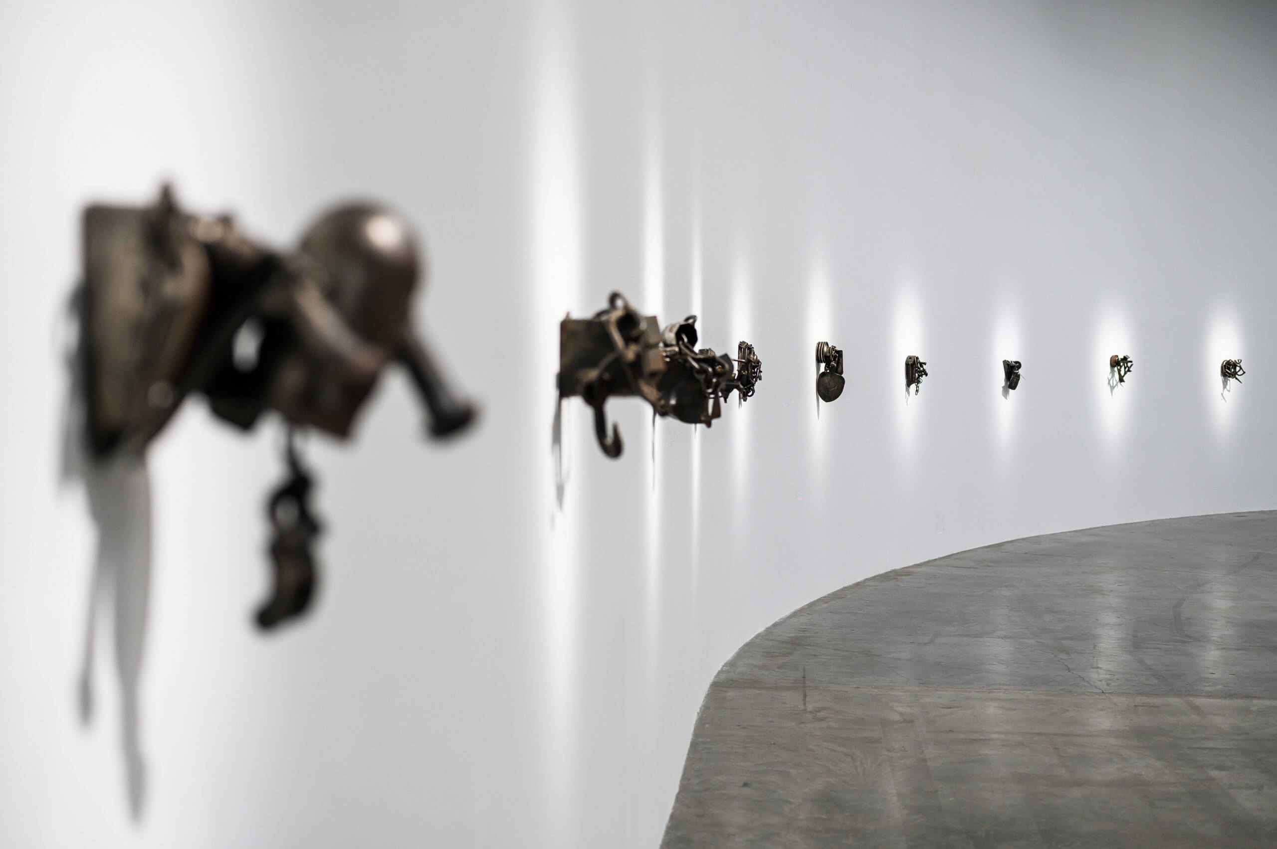 A row of small bronze sculptures mounted on a white curved gallery wall, with a polished concrete floor visible below.