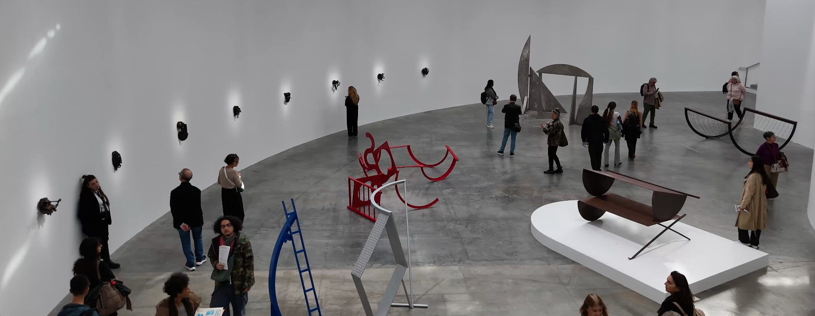 People view modern sculptures and wall-mounted art in a spacious, curved gallery with a high ceiling.