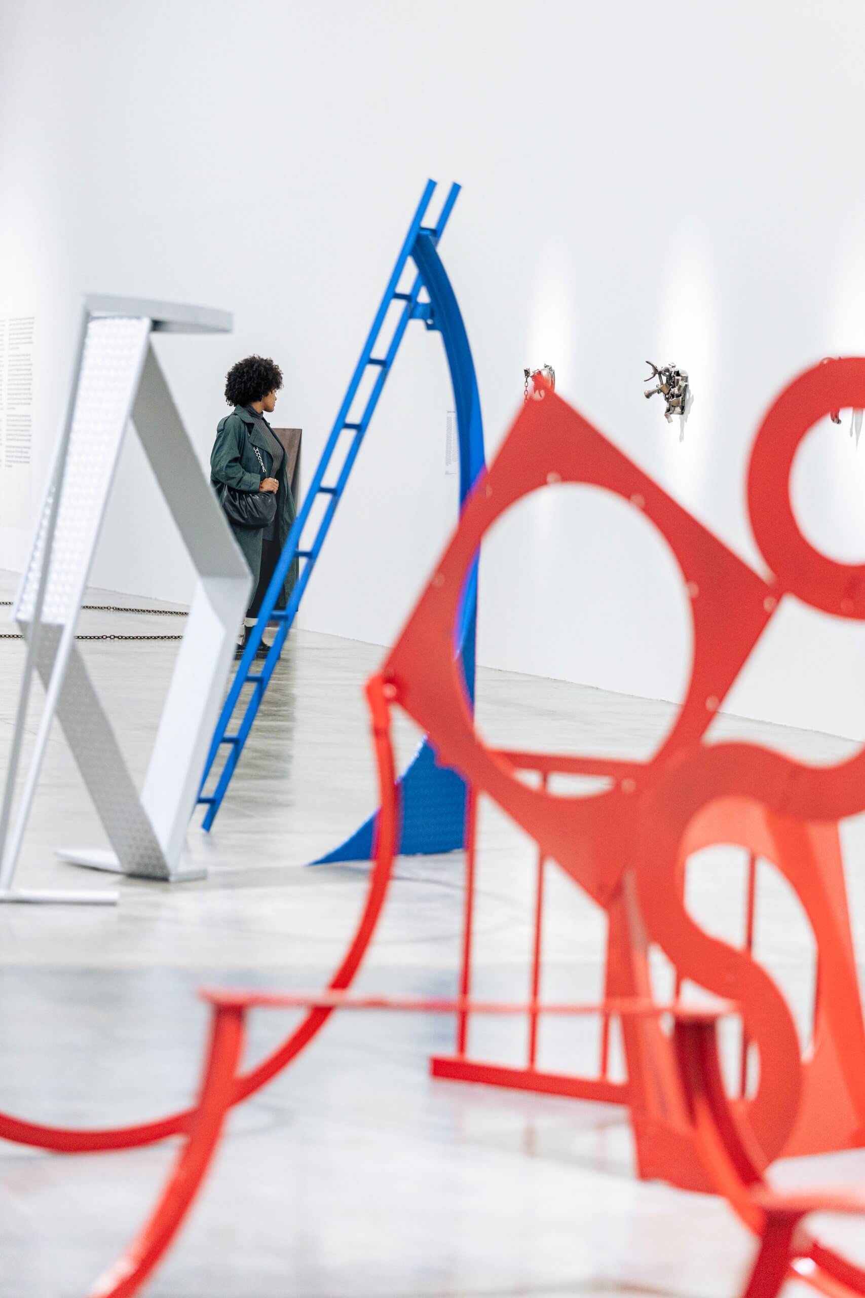 A person observes abstract metal sculptures in red, blue, and silver displayed in a modern, white-walled art gallery.