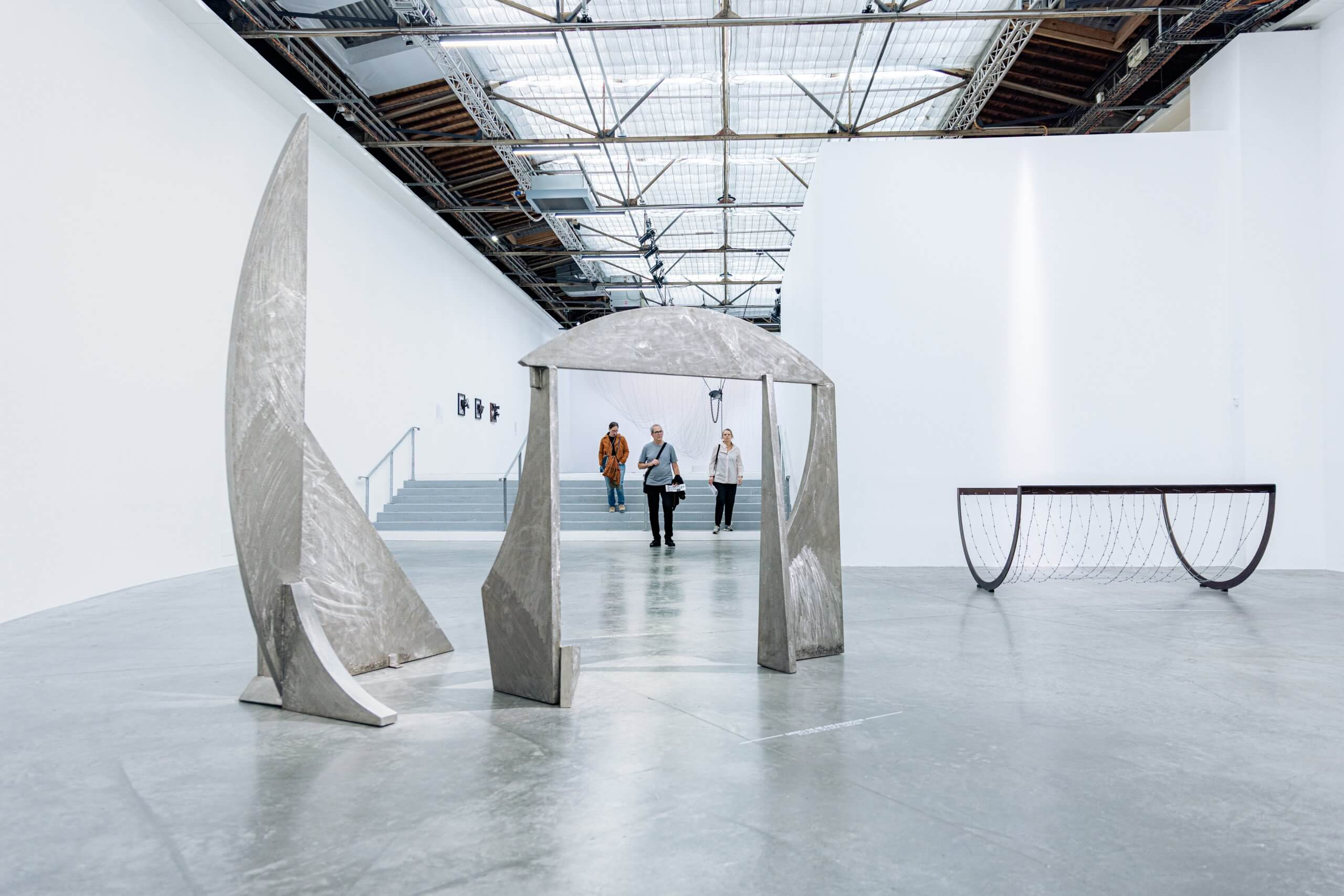 Trois personnes entrent dans une galerie d'art spacieuse où sont exposées de grandes sculptures métalliques abstraites et des éléments architecturaux modernes.