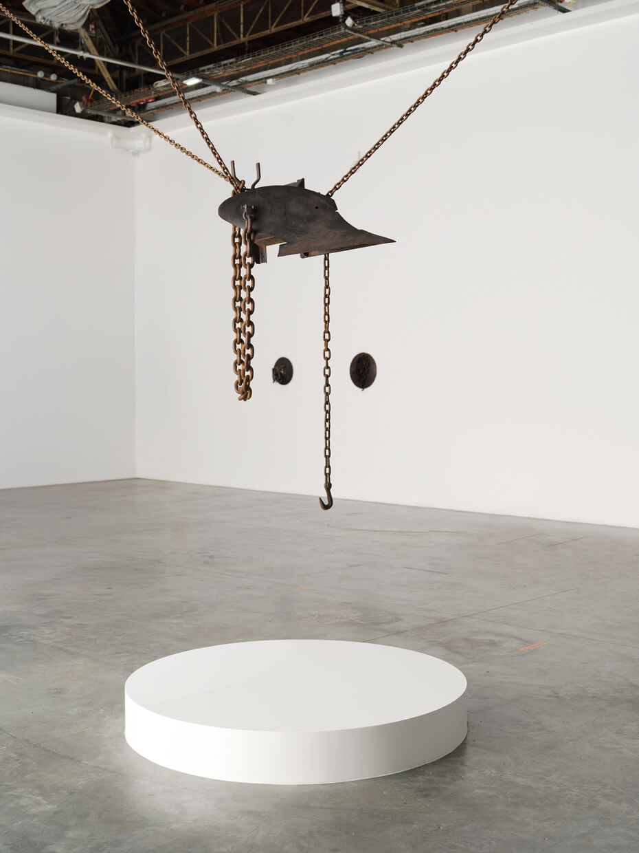 A large metal object with chains and a hook is suspended from the ceiling above a round white platform in a spacious, minimalist gallery.
