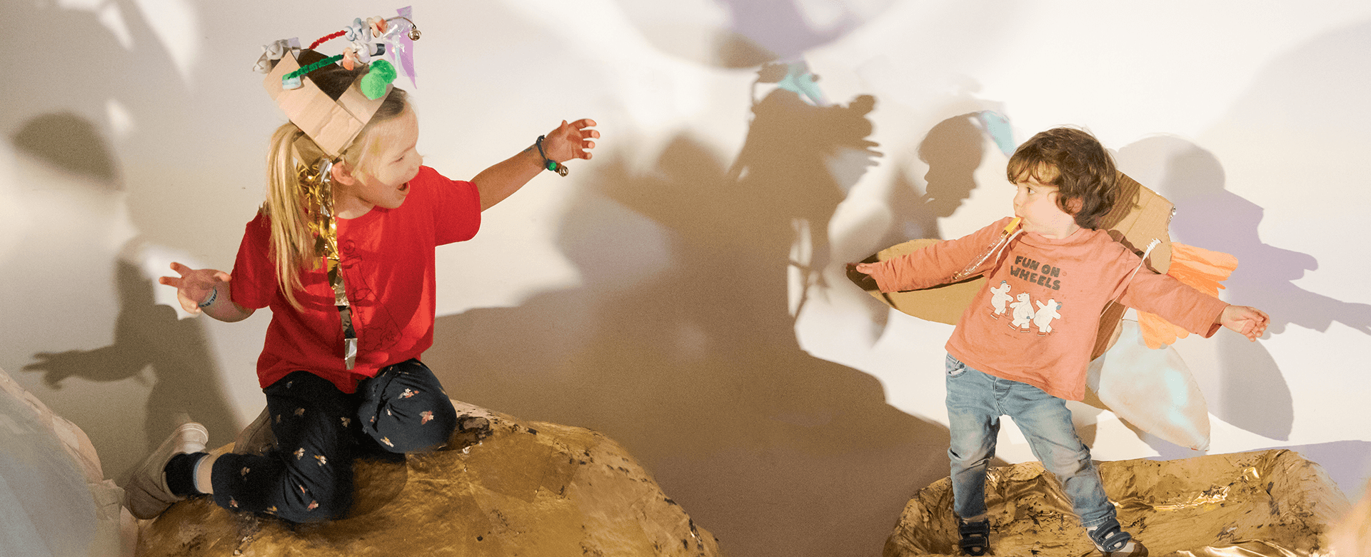 Deux enfants déguisés se font face, bras tendus, debout sur des rochers. Leurs ombres sont projetées sur le mur derrière eux.