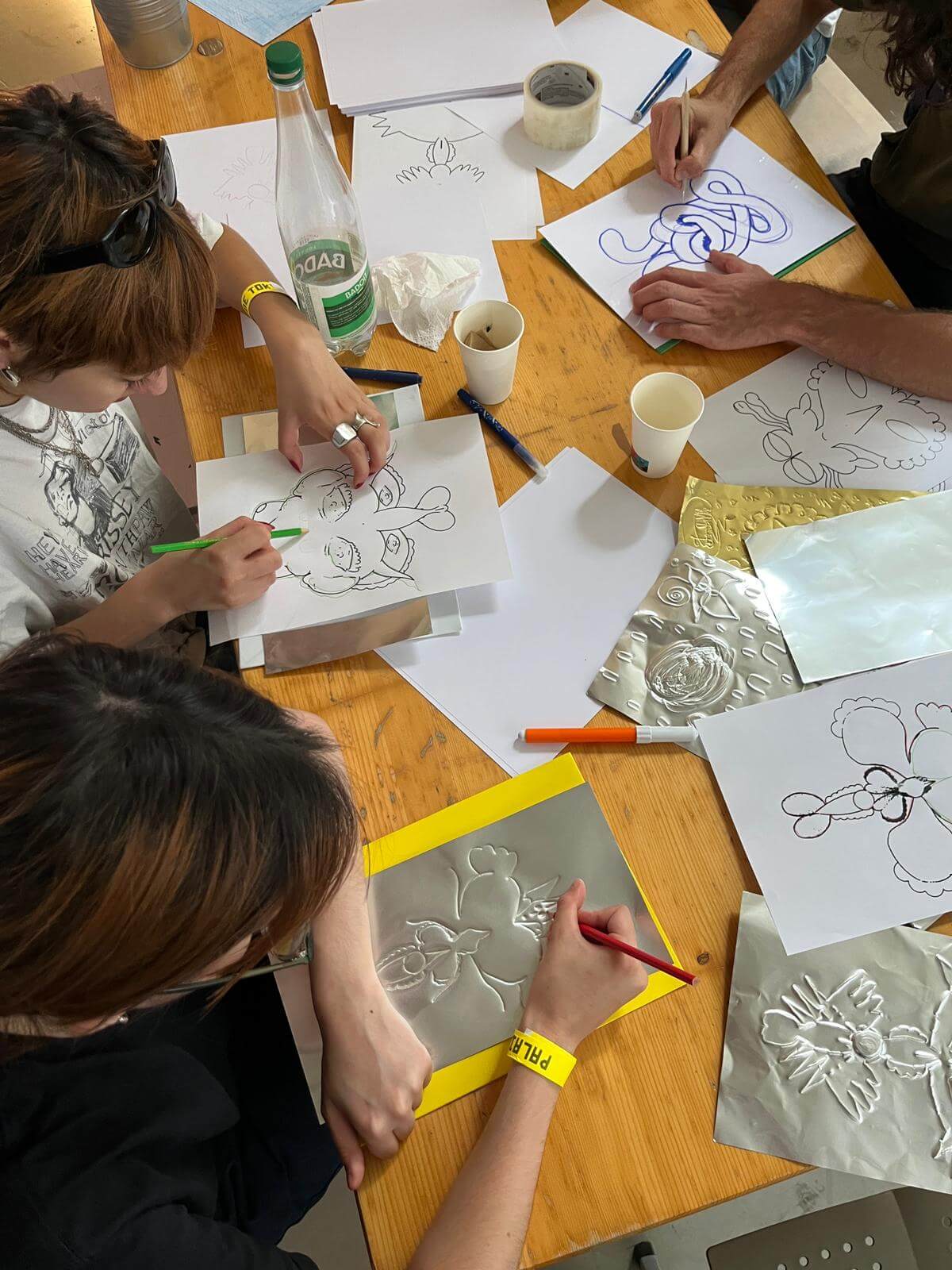 Trois personnes sont assises à une table et dessinent des motifs floraux sur du papier et des feuilles de métal, entourées de fournitures artistiques, de tasses et d'une bouteille d'eau.
