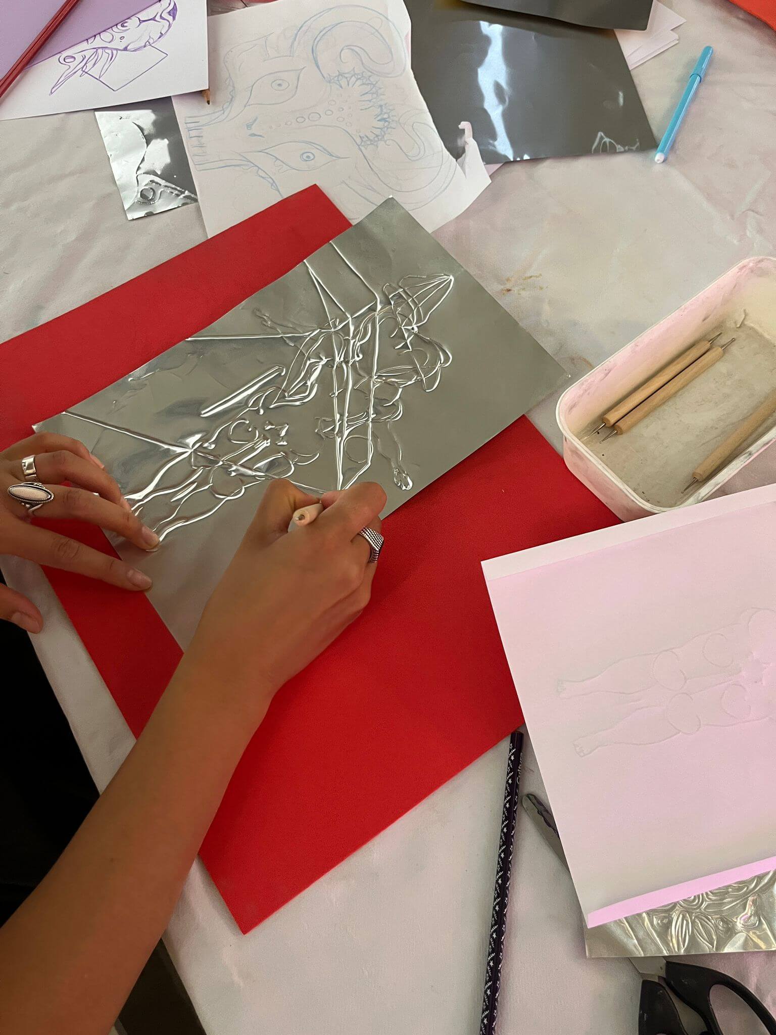 Une personne trace un dessin sur une feuille de métal à l'aide d'un stylet, les croquis de référence et les outils étant visibles sur la table.