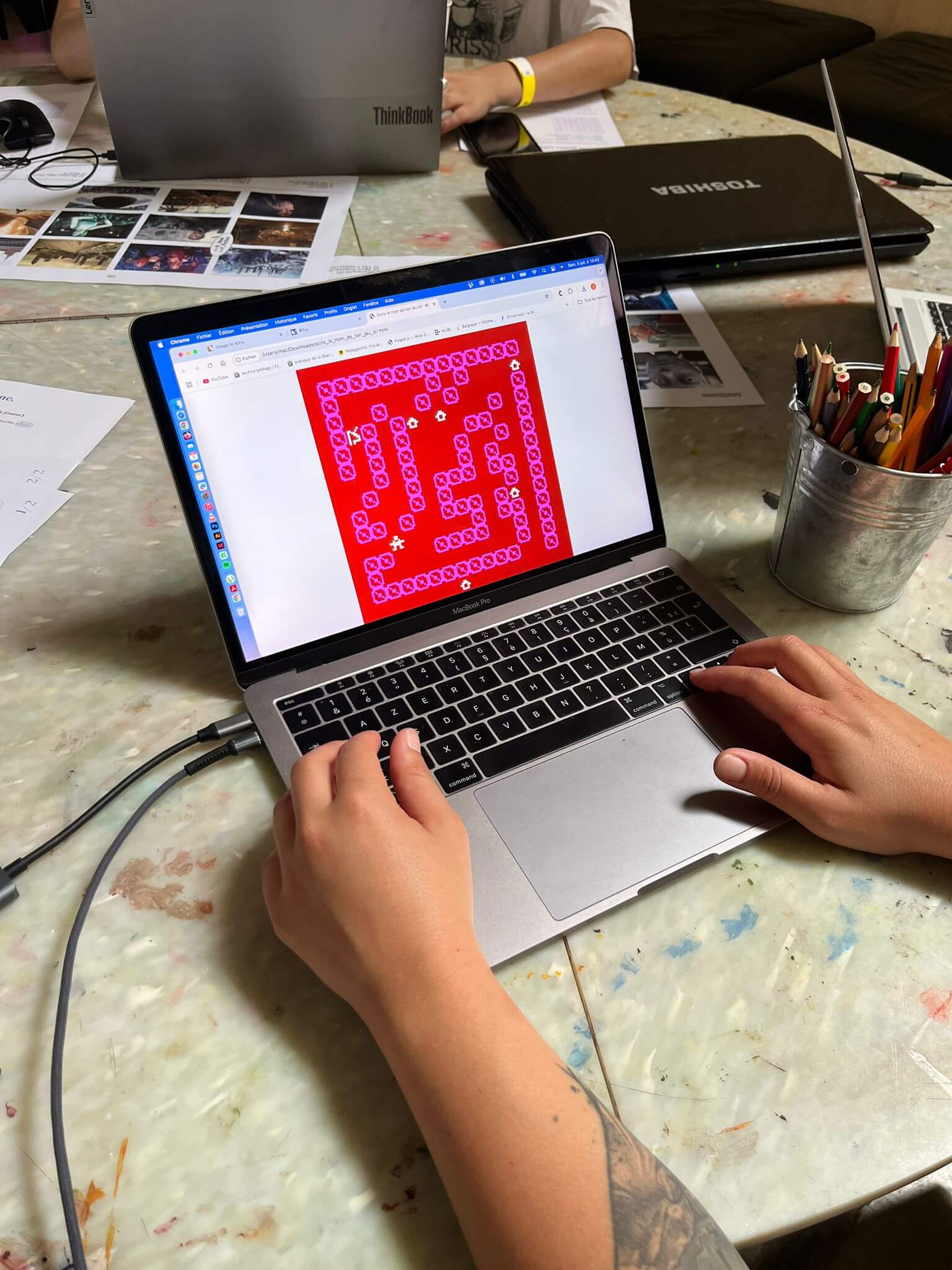 Personne utilisant un ordinateur portable affichant un motif géométrique rouge et rose ; des fournitures artistiques et des papiers sont posés sur la table dans un espace de travail commun.