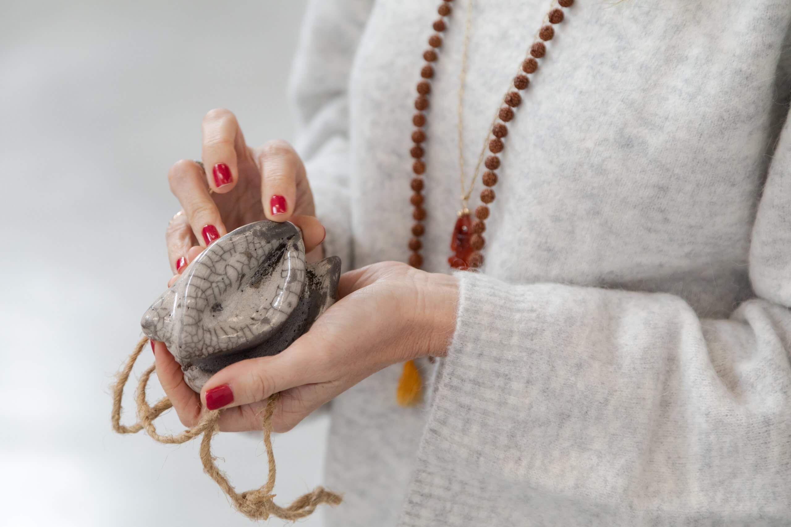 Une personne vêtue d'un pull-over gris tient un objet en pierre sculptée avec une corde. Elle porte du vernis à ongles rouge et un collier de perles.
