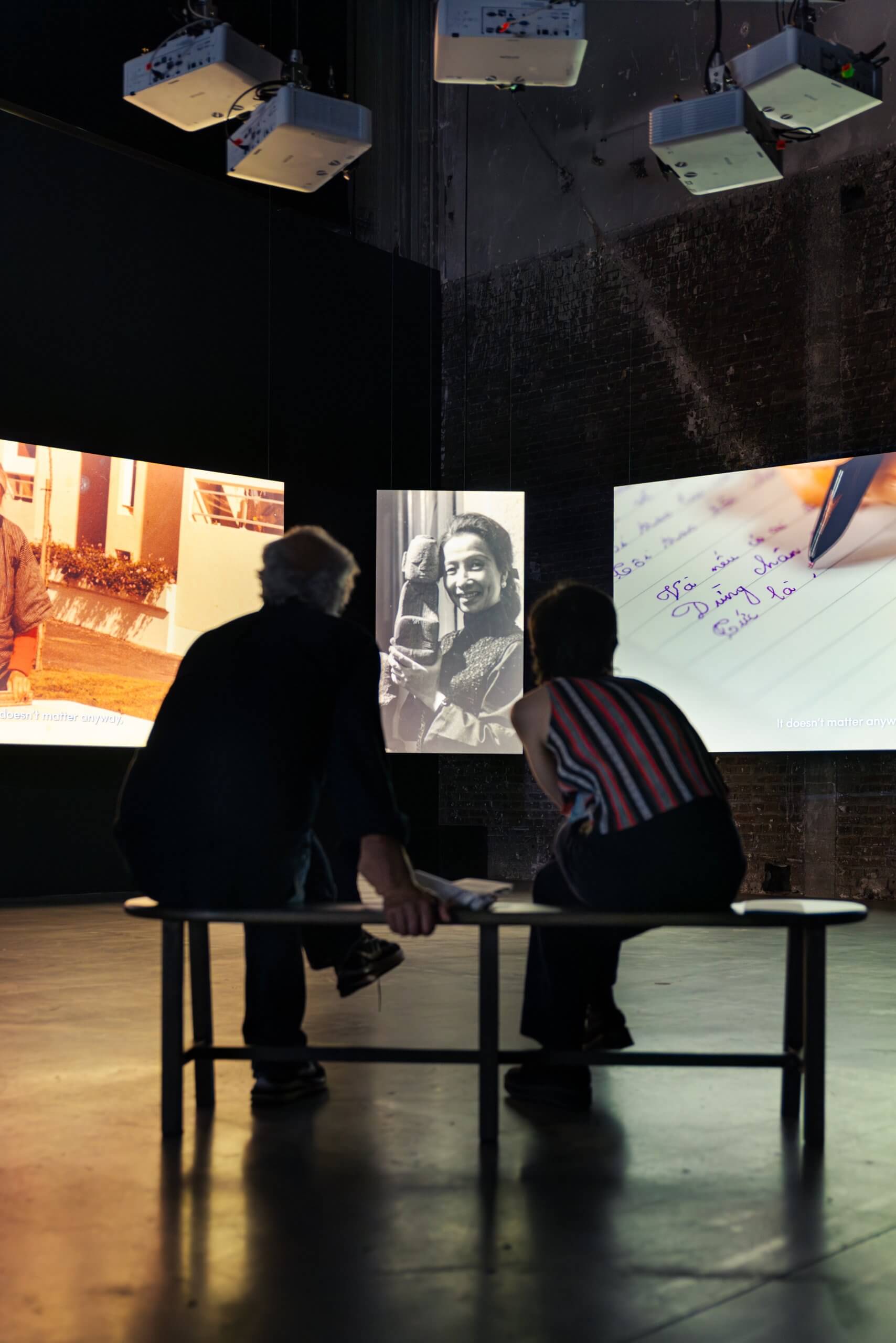 Deux personnes sont assises sur un banc dans une pièce faiblement éclairée, et regardent de grandes images projetées de vieilles photographies et de textes manuscrits sur des écrans environnants.