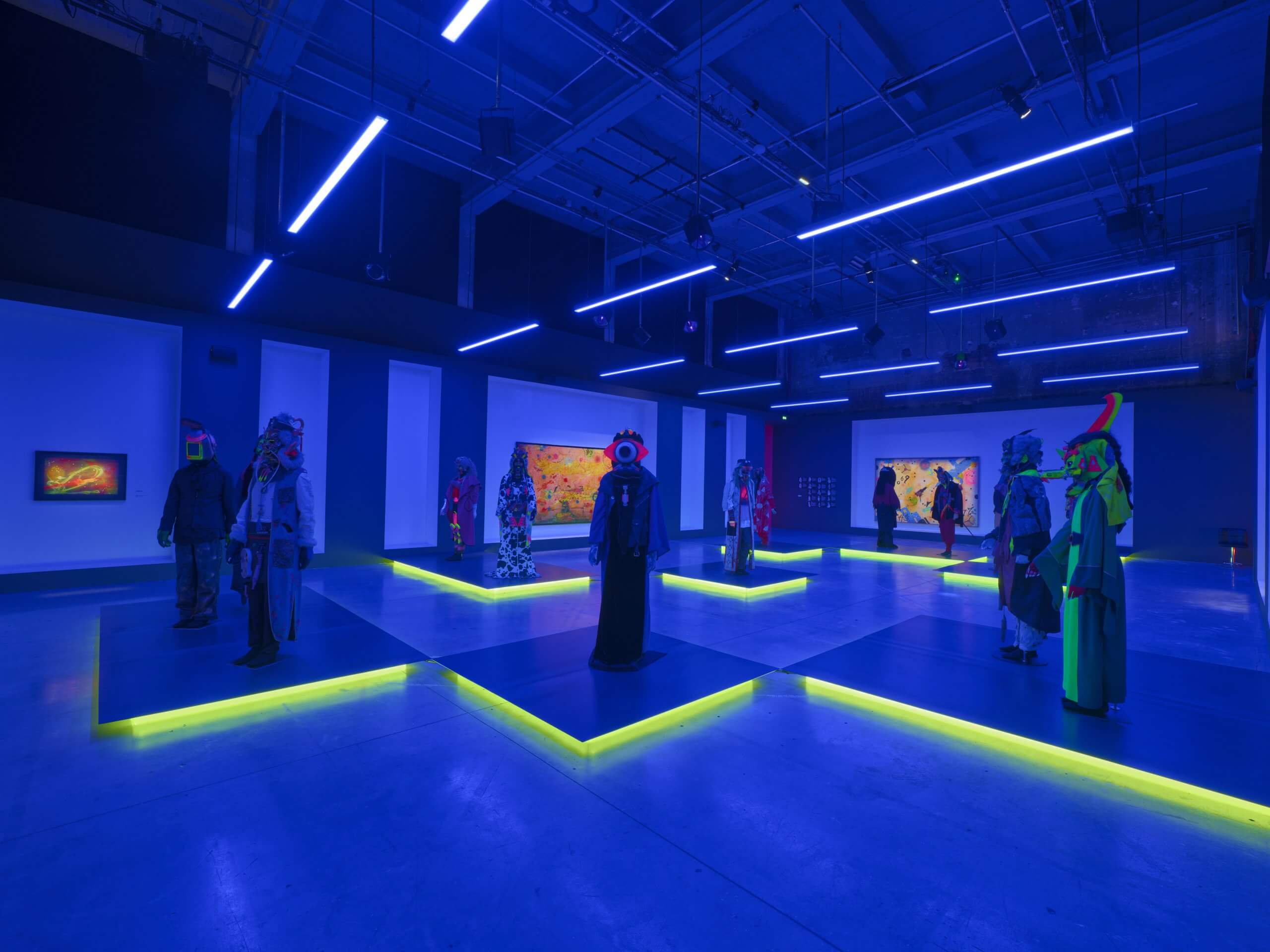 Des personnes portant des costumes et des masques colorés se tiennent à distance dans une galerie faiblement éclairée, aux tons bleus, avec des lampes de sol jaune néon et des œuvres d'art illuminées sur les murs.