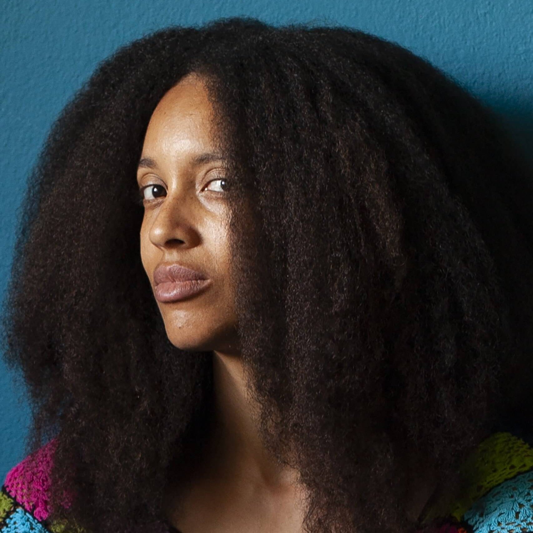 Une personne aux longs et volumineux cheveux bouclés, vêtue d'un haut en crochet coloré, est assise devant un mur bleu et regarde légèrement sur le côté.