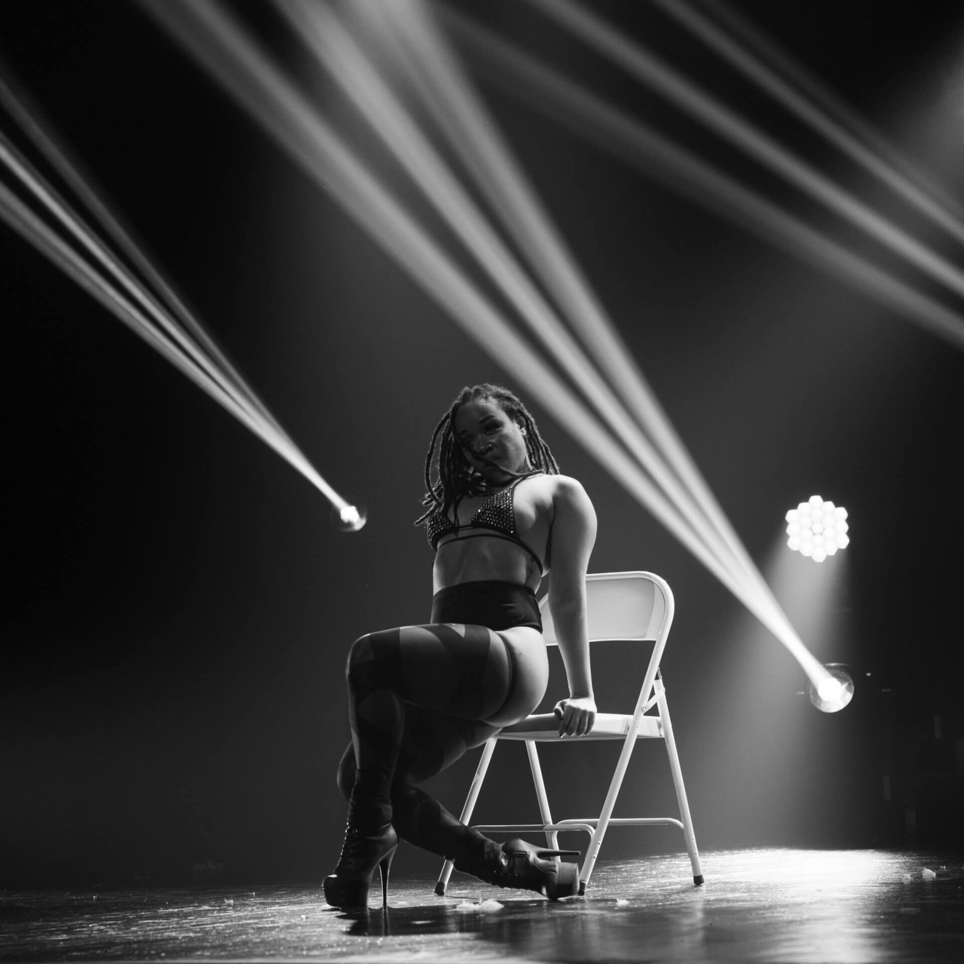 Une personne en lingerie et talons hauts est assise sur une chaise pliante, les jambes croisées, sous des lumières de scène dont les faisceaux sont visibles dans un théâtre sombre.