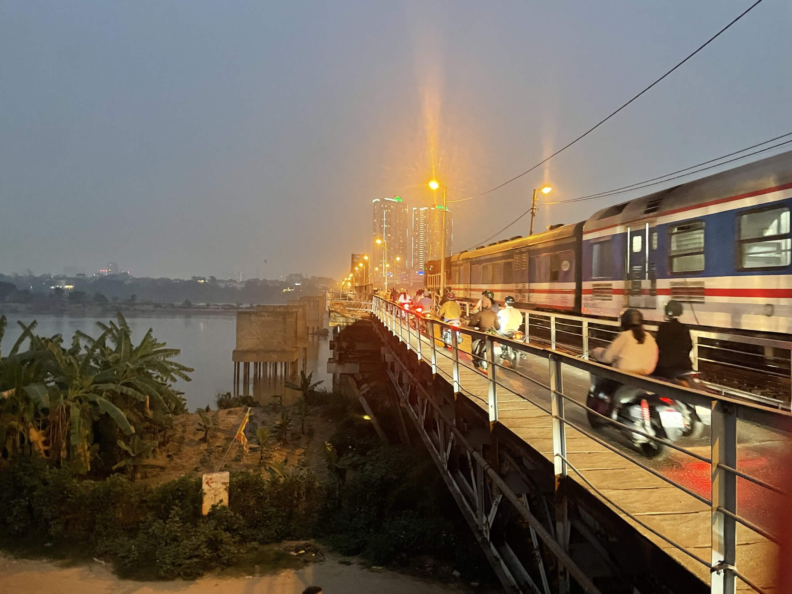 Un train passe à côté de motos sur un pont au crépuscule, avec les lumières de la ville et les grands bâtiments visibles au loin.