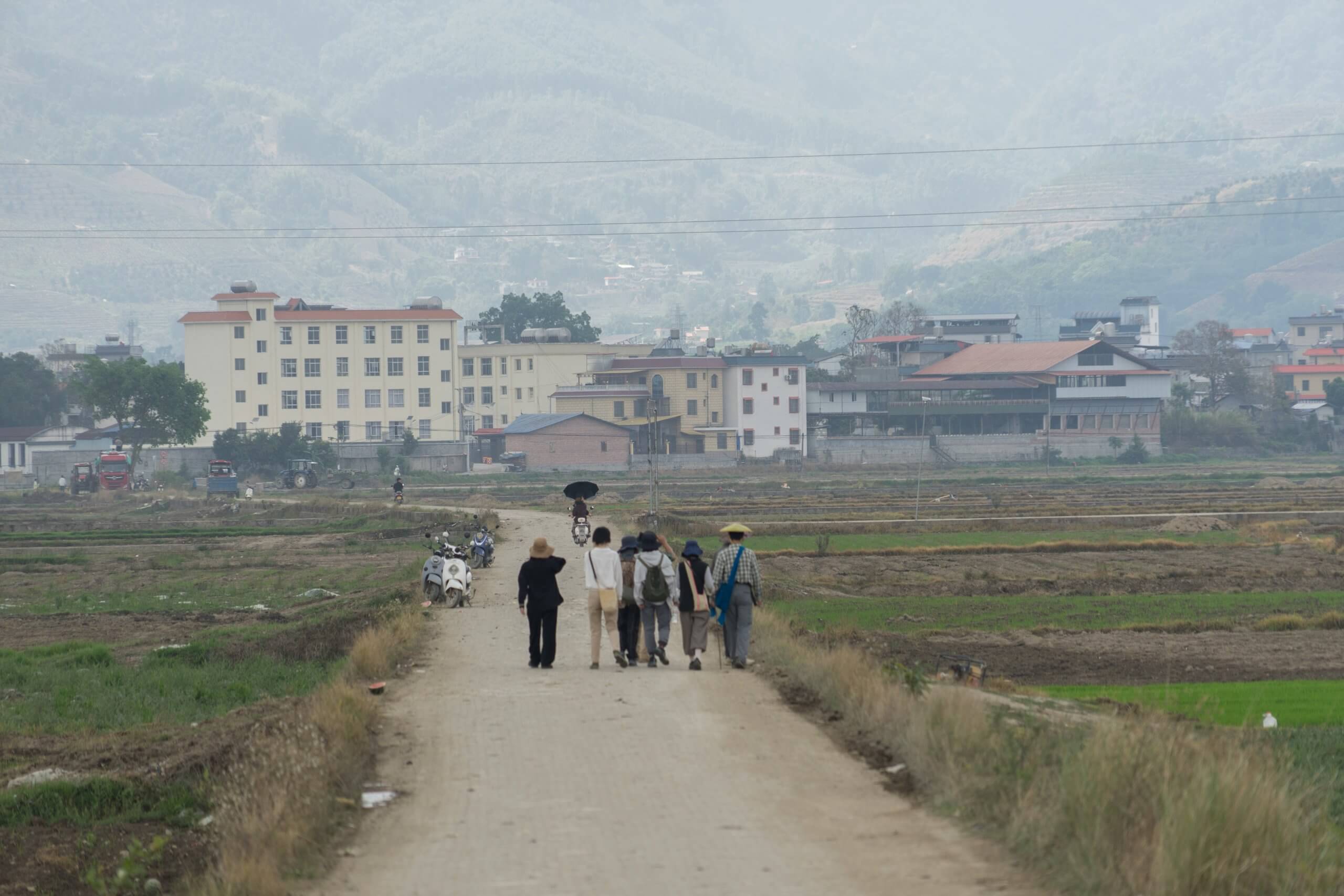 Un groupe de personnes marche sur un chemin de terre à travers des champs en direction d'une ville avec des bâtiments et des montagnes en arrière-plan.