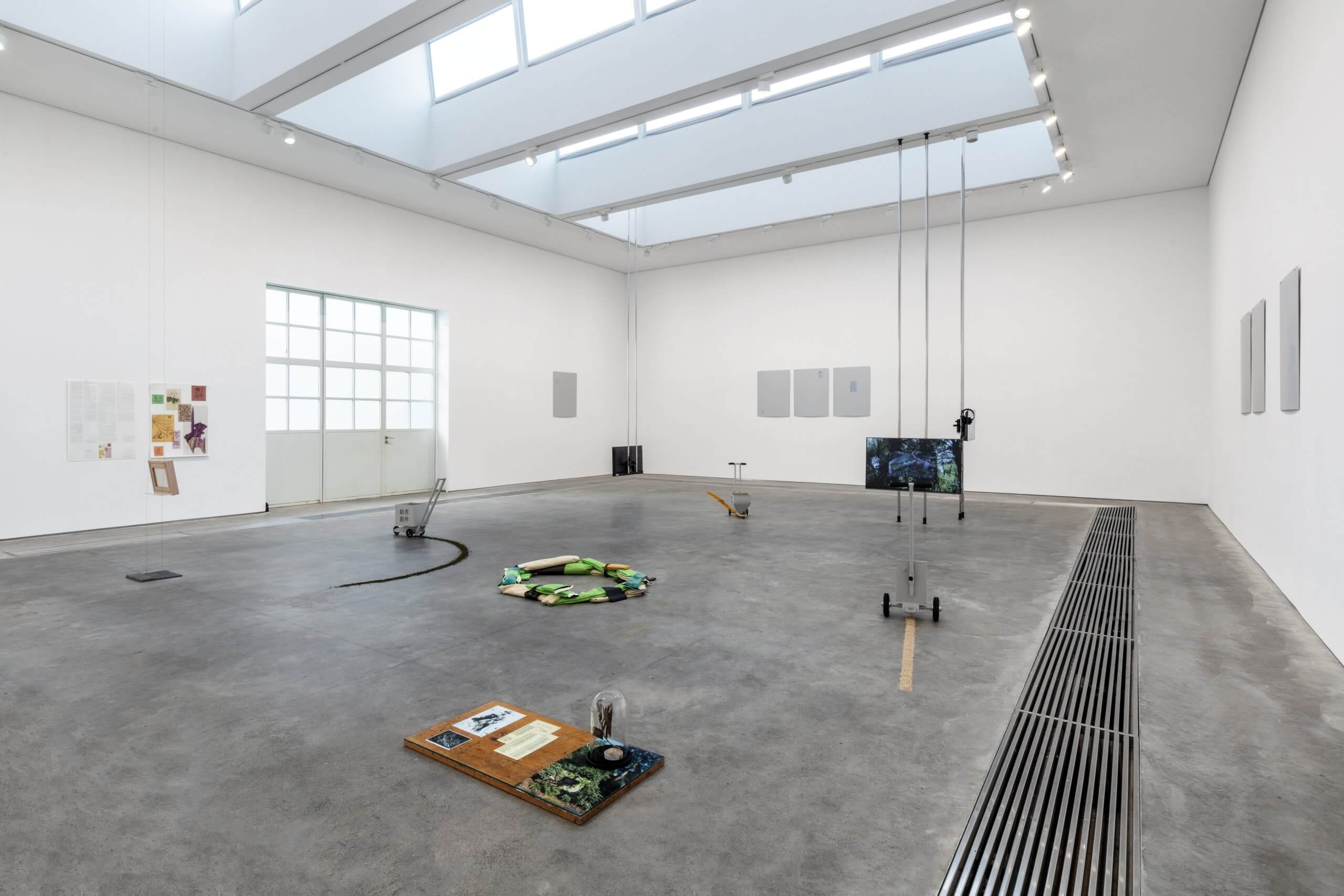 Une galerie d'art contemporain avec diverses installations mixtes placées sur le sol et des œuvres minimales encadrées sur des murs blancs sous un éclairage naturel.