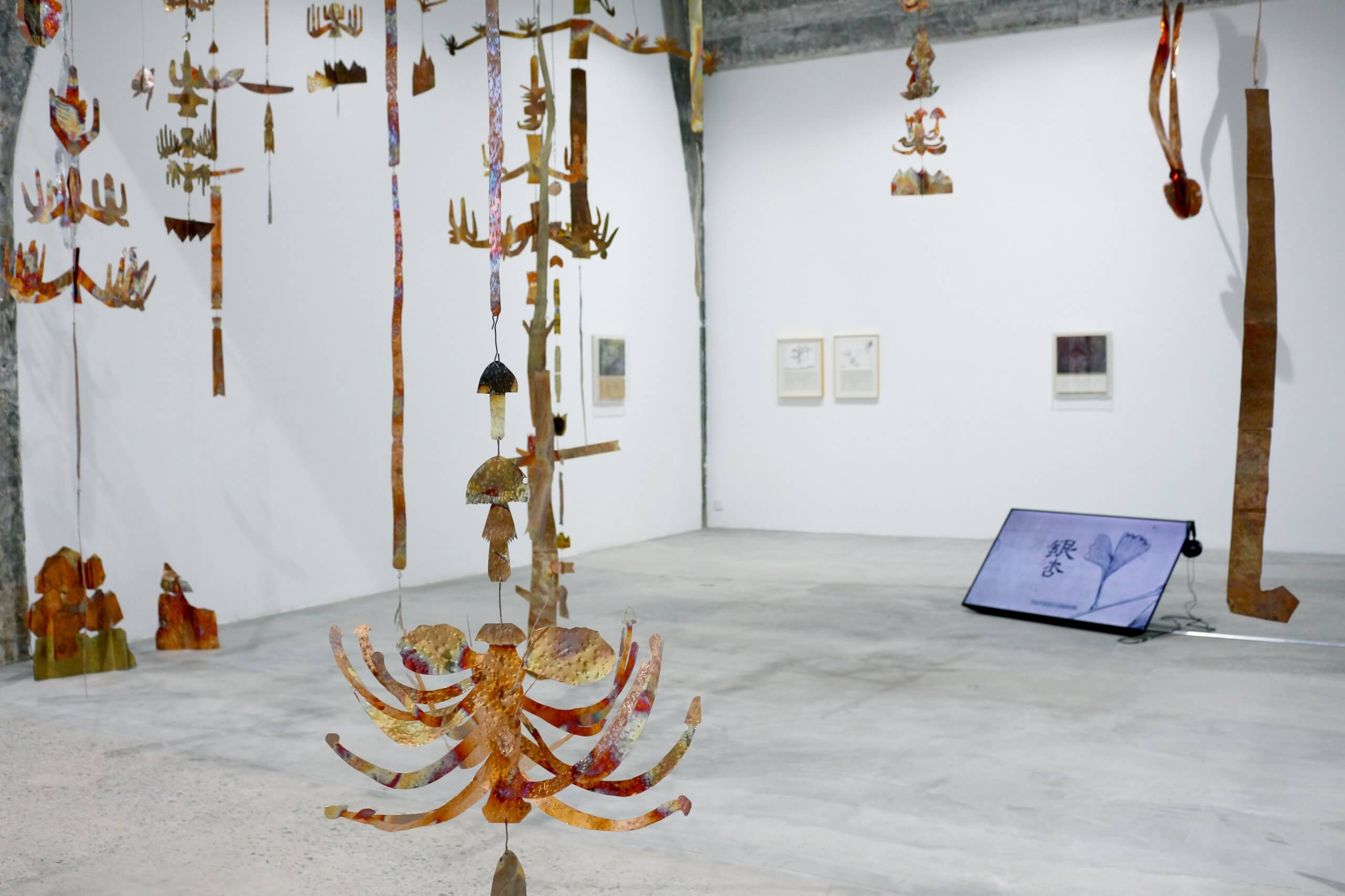 Une galerie d'art contemporain présente des sculptures abstraites suspendues, des dessins encadrés sur des murs blancs et un écran numérique au sol.