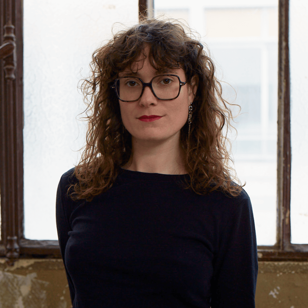 Une personne aux cheveux bruns bouclés, portant des lunettes et un haut noir se tient à l'intérieur devant des fenêtres givrées, regardant la caméra avec une expression neutre.