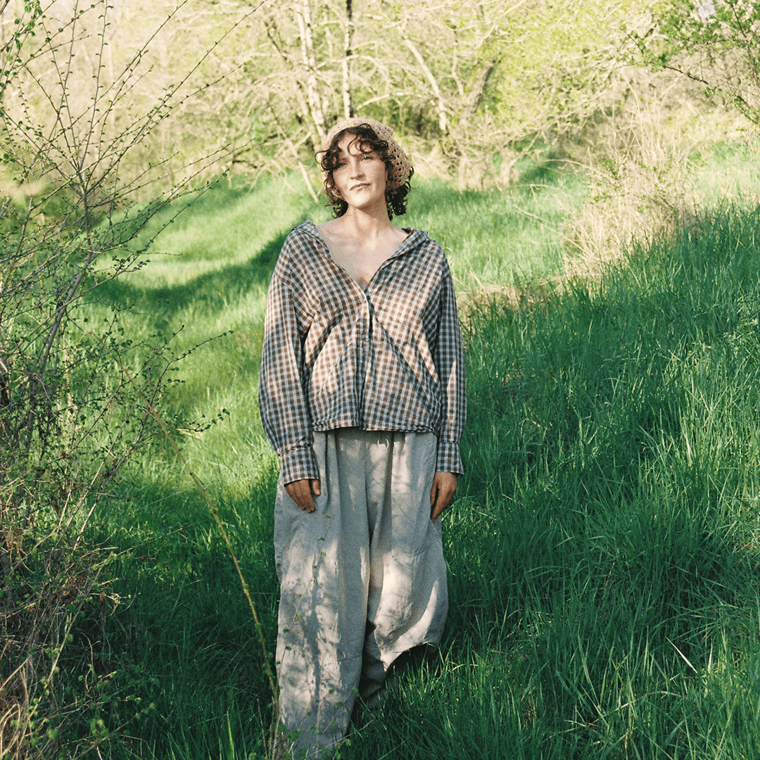 Une personne vêtue d'une chemise à carreaux et d'un pantalon ample se tient dans une zone herbeuse et ensoleillée, avec des arbres et des buissons en arrière-plan.