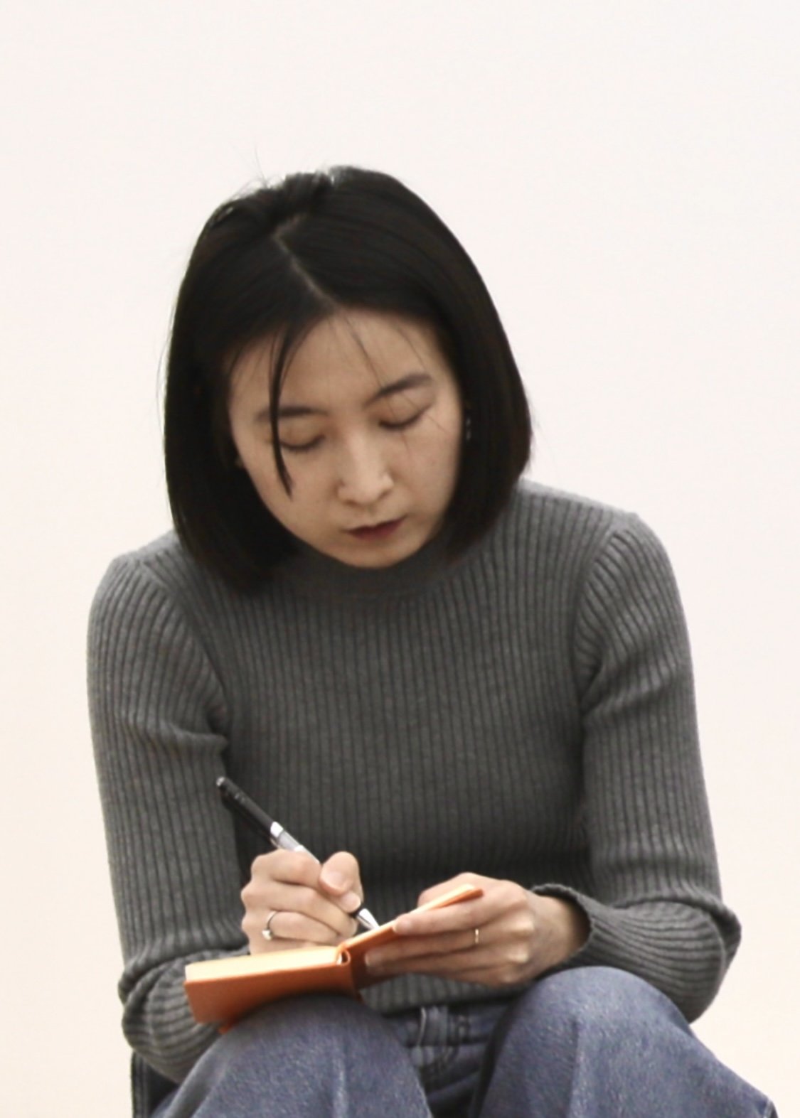 Une personne aux cheveux noirs mi-longs est assise et écrit dans un carnet orange avec un stylo. Elle porte un pull gris côtelé et un jean bleu.