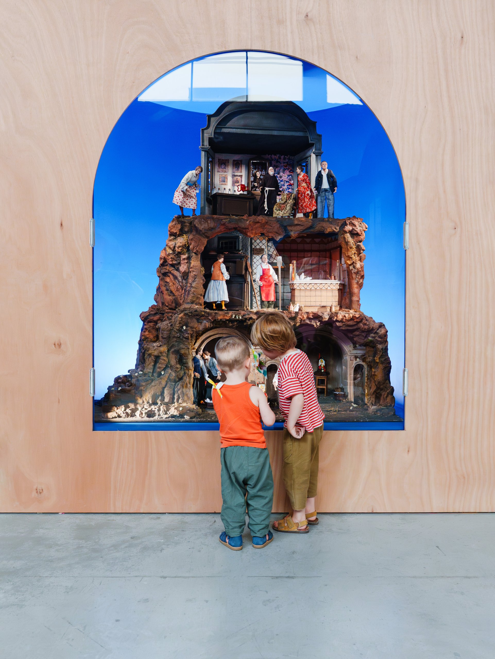 Deux jeunes enfants se tiennent devant une vitrine en bois contenant un diorama miniature très détaillé, à plusieurs niveaux, avec de petites figurines humaines et un fond bleu.