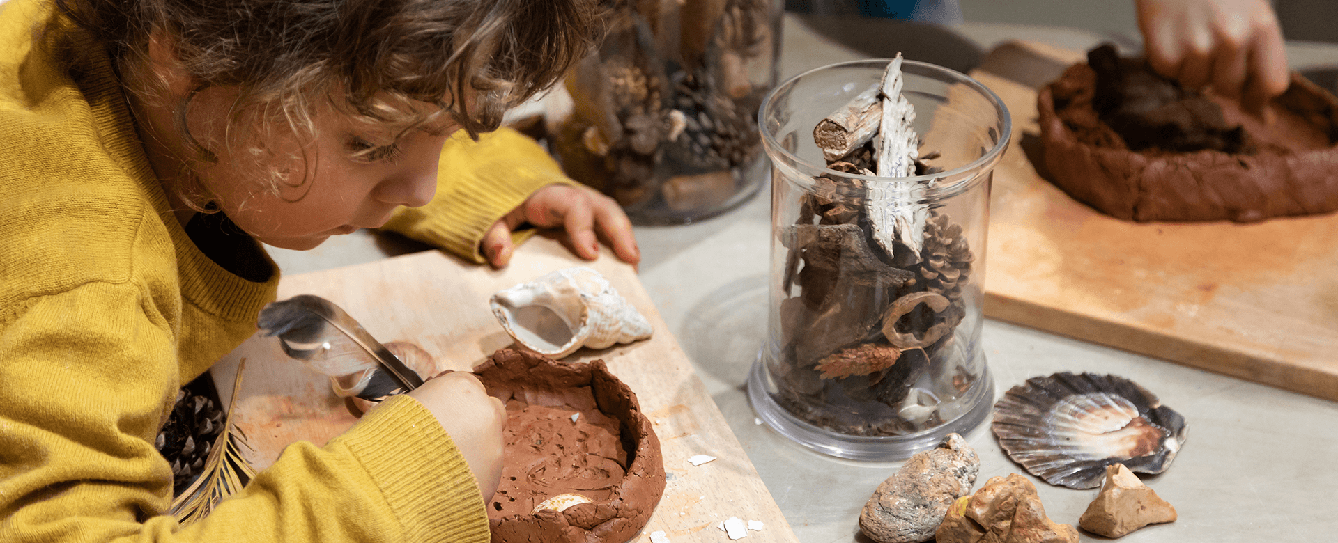Un enfant vêtu d'un pull jaune décore de l'argile avec des objets naturels sur une table, avec des bocaux de coquillages, de plumes et de rochers à proximité.