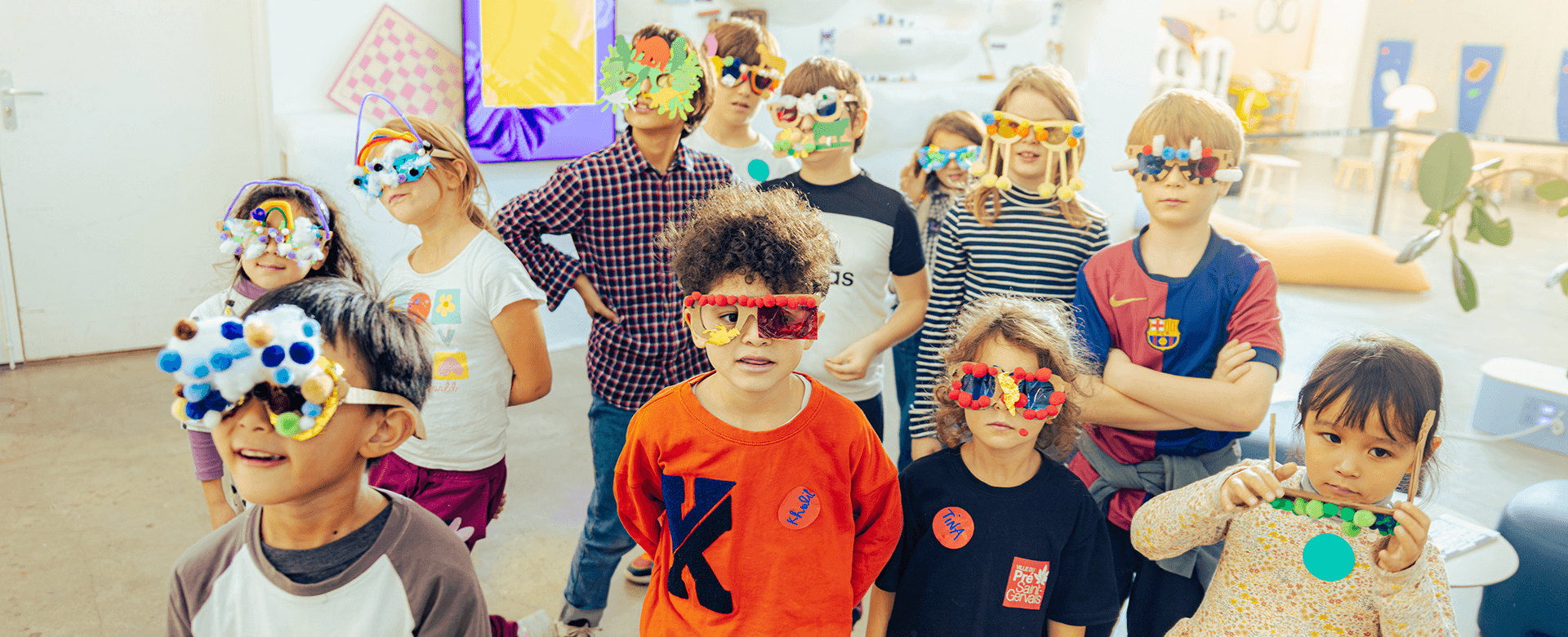Un groupe d'enfants portant des lunettes colorées, fabriquées à la main, se tient ensemble dans une salle de classe ou de jeu bien éclairée.