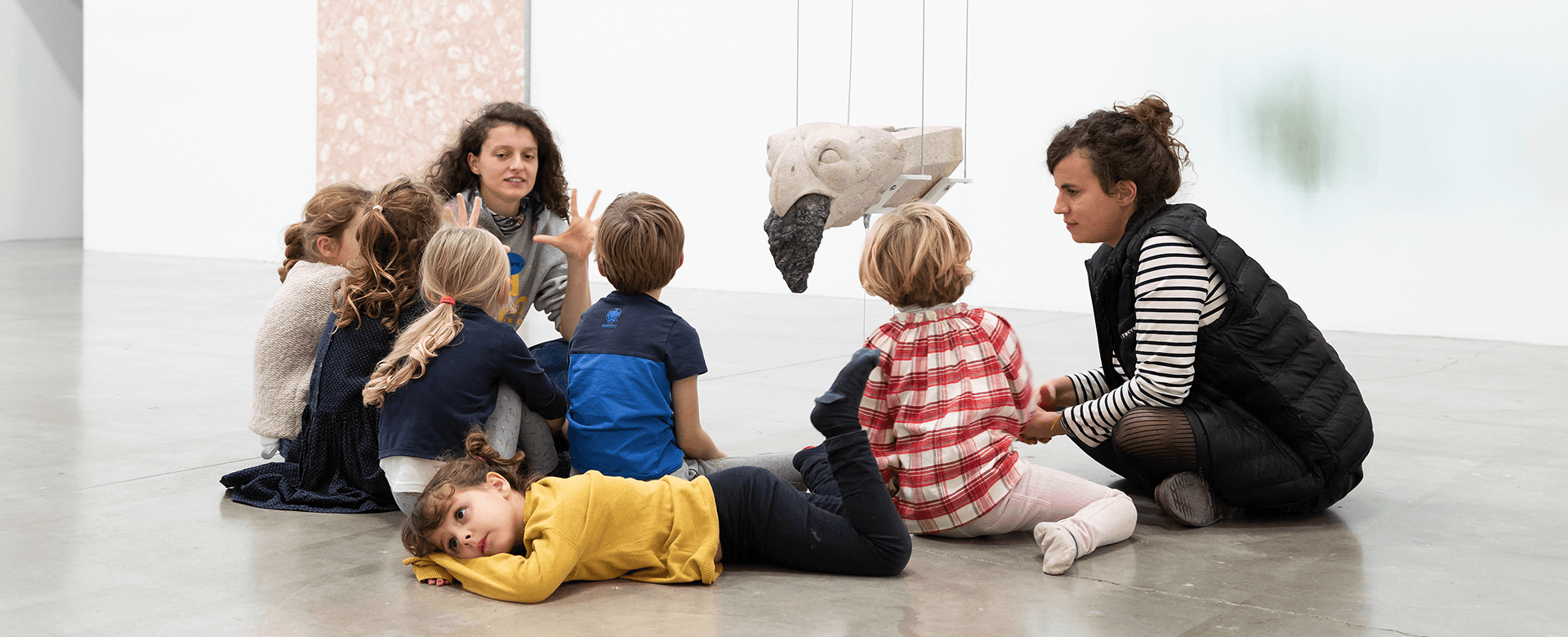 Un groupe d'enfants est assis par terre avec deux adultes dans une galerie, écoutant et regardant une sculpture suspendue.