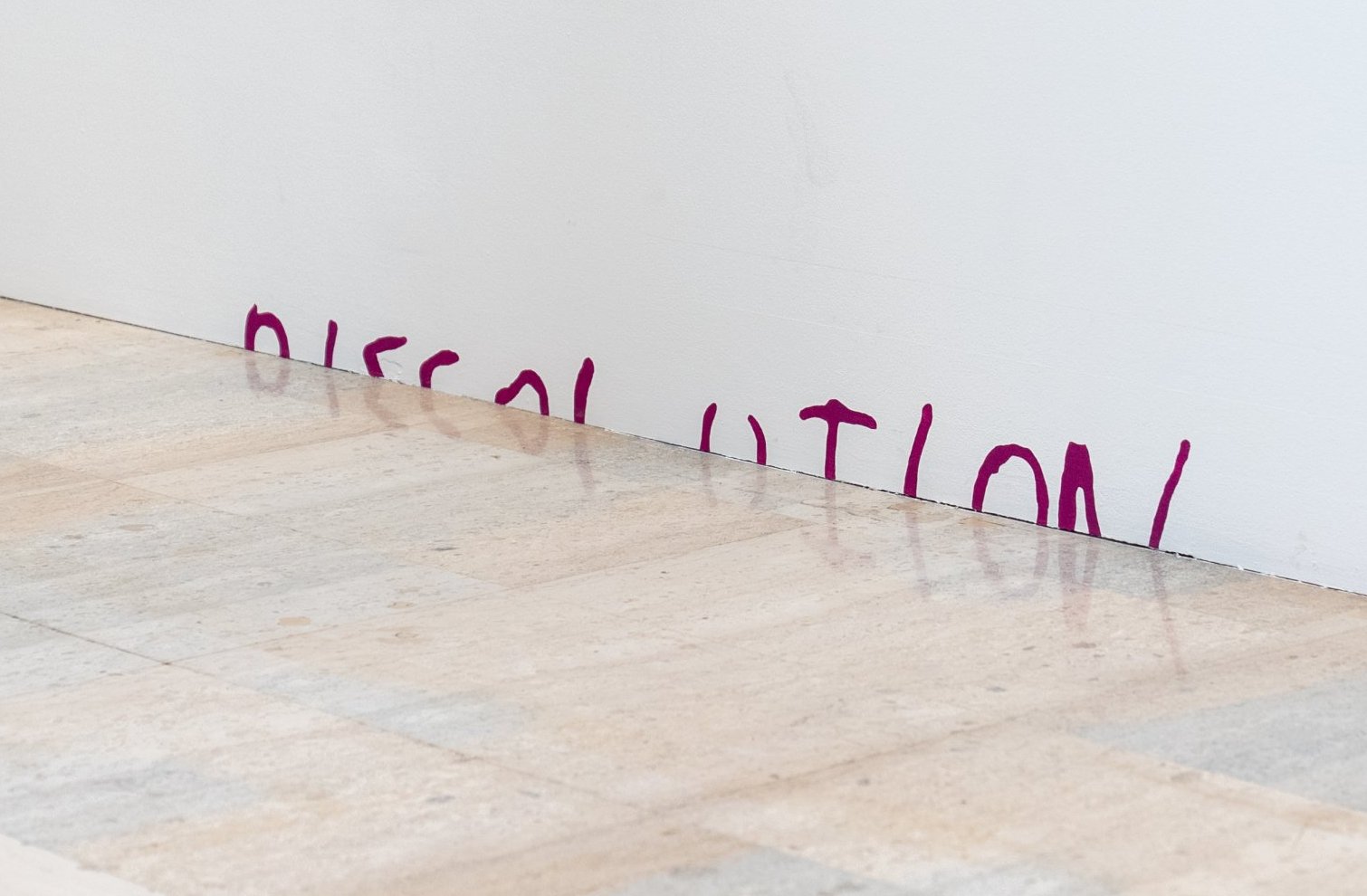 Un graffiti violet sur un mur blanc montre un mot partiellement visible, DISSOLUTION, et des dessins au trait simples, avec un sol carrelé de couleur beige clair au premier plan.