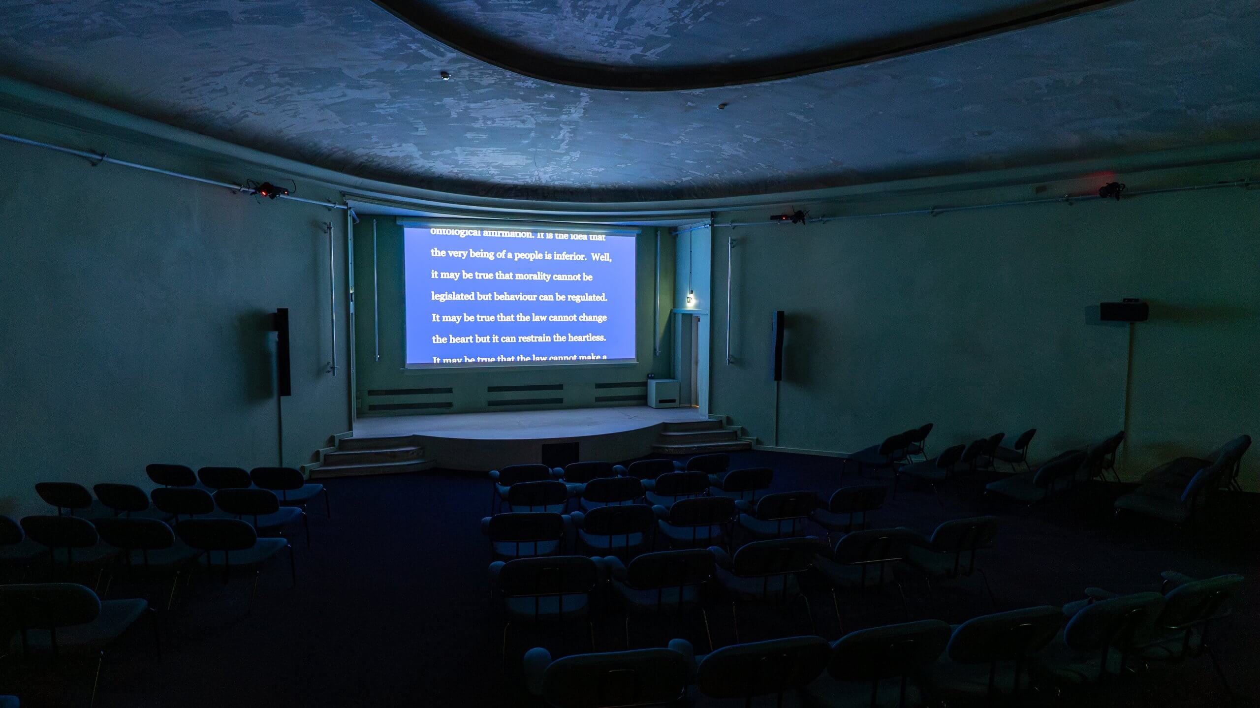 Un auditorium vide avec des rangées de chaises fait face à un grand écran affichant un texte blanc sur fond bleu. La salle est faiblement éclairée par des lampes au plafond et au sol.
