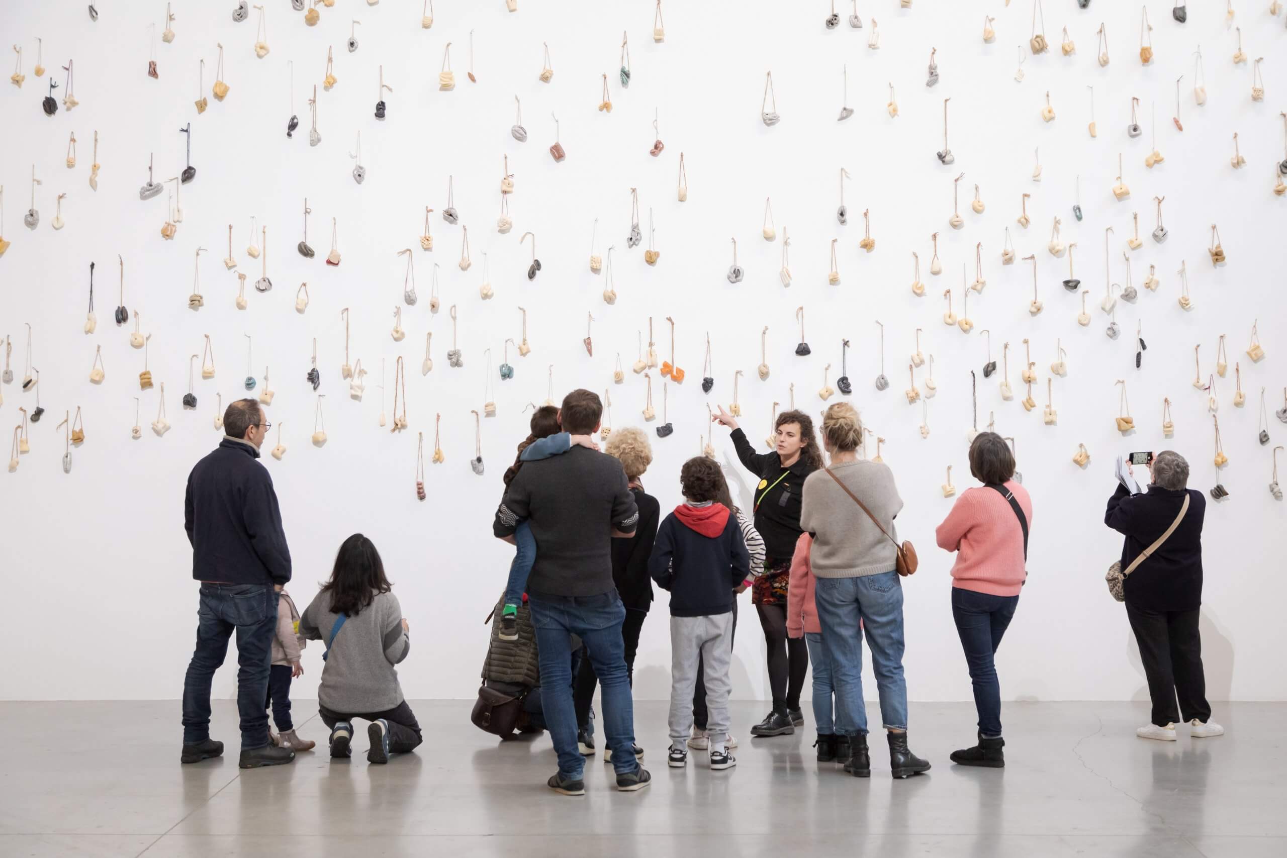 Un groupe de personnes observe un mur de galerie blanc recouvert de nombreux objets suspendus, guidé par une personne faisant des gestes en direction de l'installation artistique.