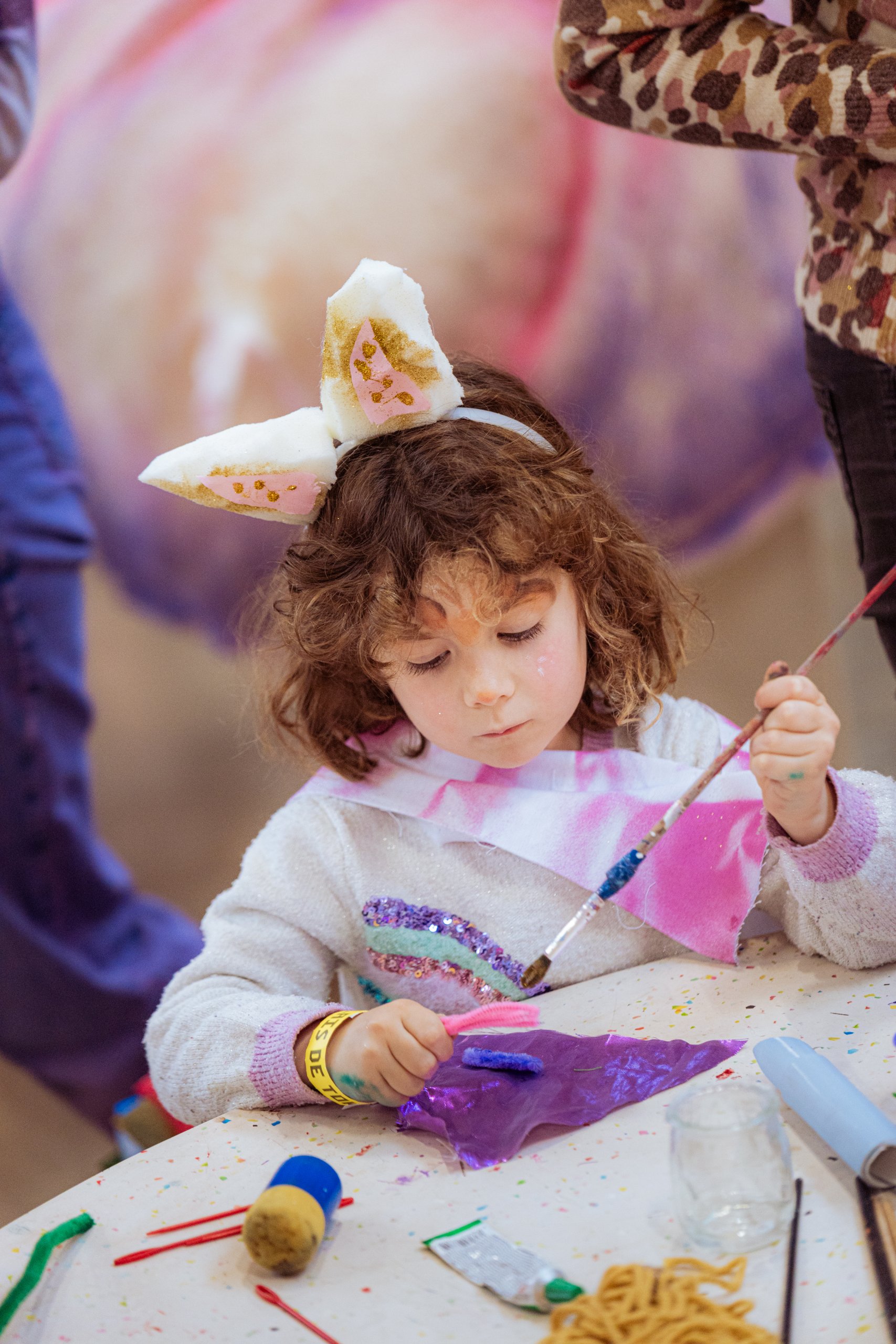 Un jeune enfant portant des oreilles de lapin peint un objet violet à une table de loisirs créatifs, entouré de matériel artistique, en présence d'adultes.