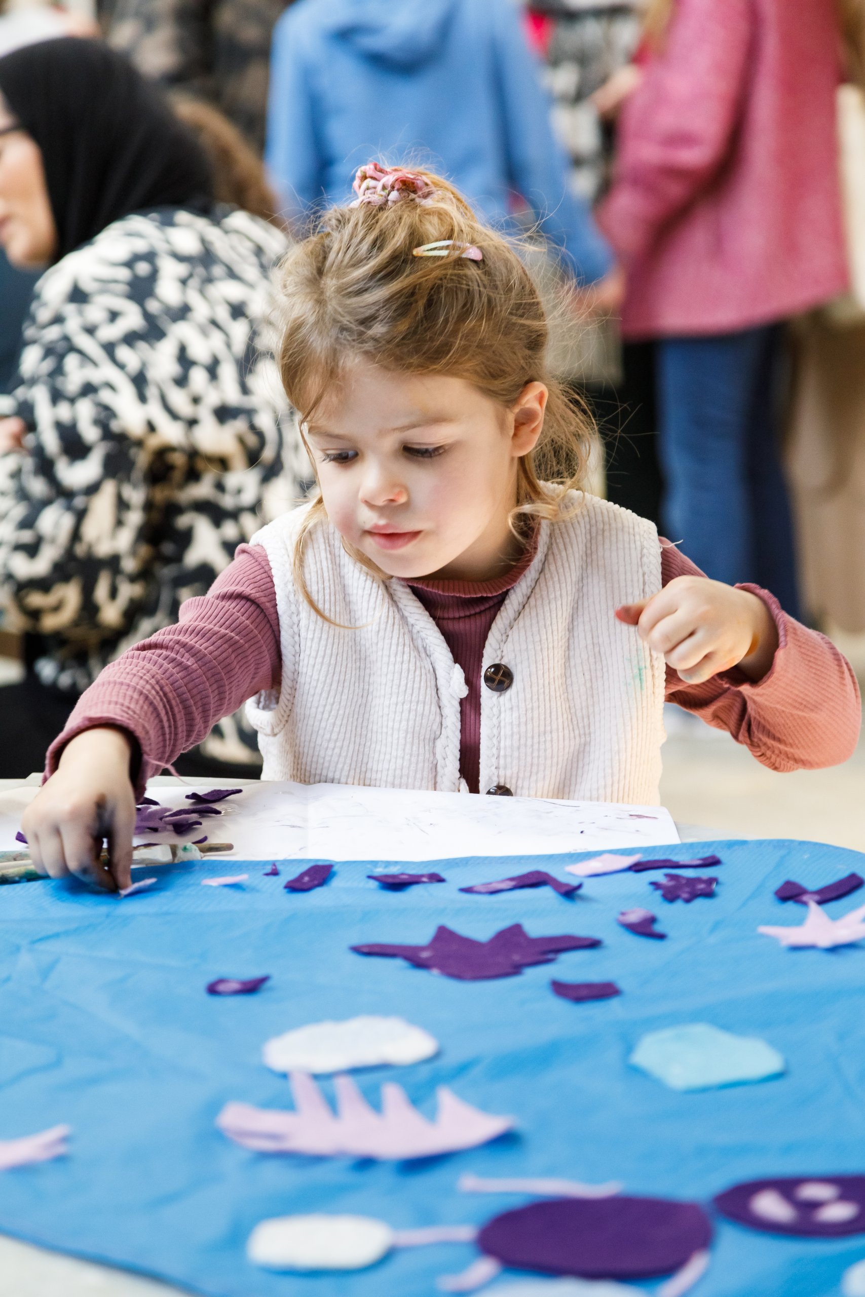 Une jeune fille est assise à une table, concentrée sur la création d'une œuvre d'art à partir de formes en papier bleu et violet. On aperçoit d'autres personnes à l'arrière-plan.