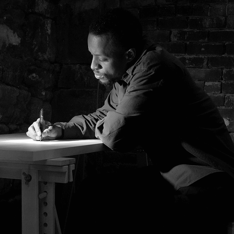 Un homme est assis à une petite table, écrivant sur un papier avec un stylo dans une pièce faiblement éclairée aux murs de briques.