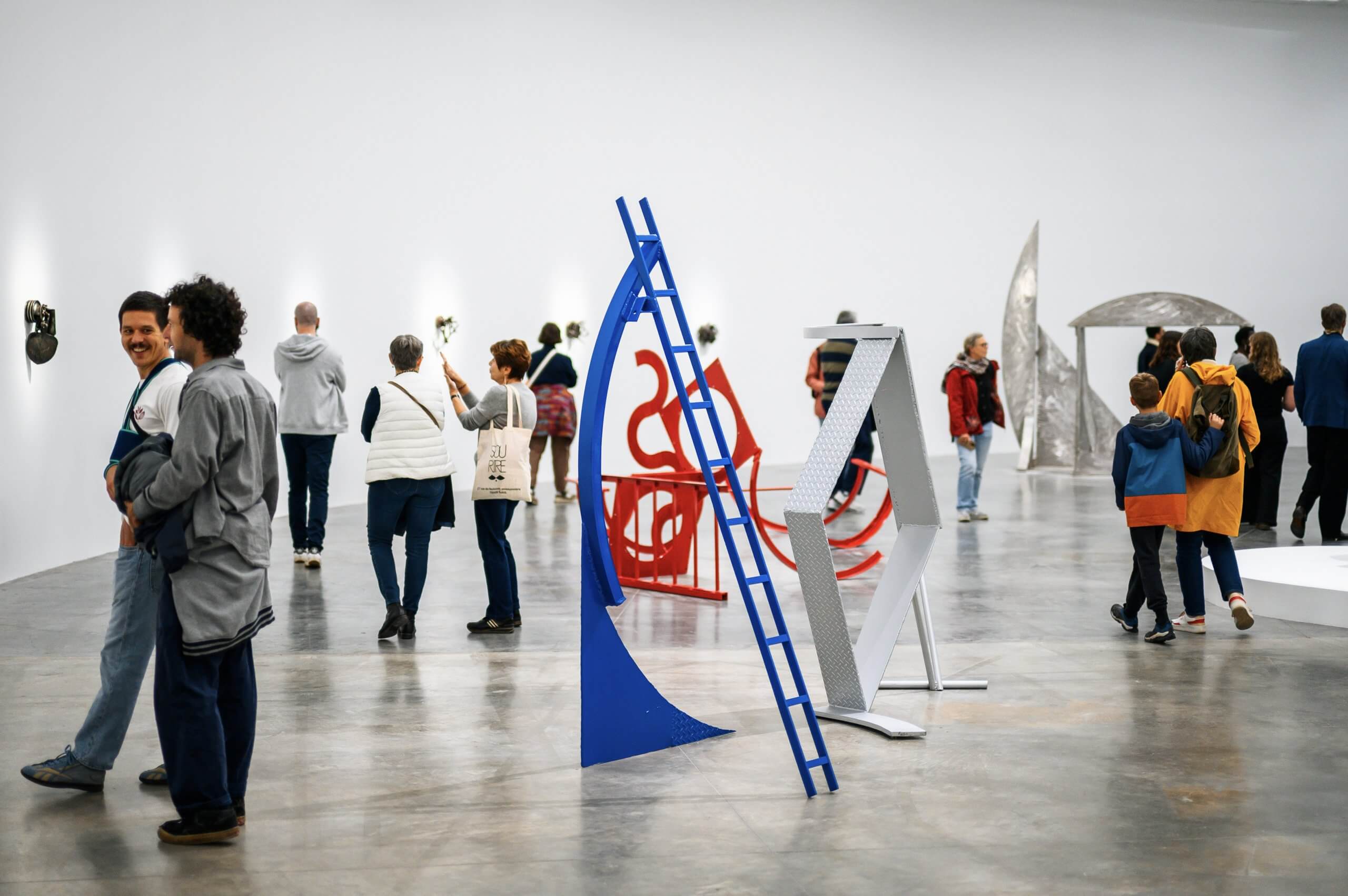 Des personnes se promènent dans une galerie d'art contemporain où de grandes sculptures et installations abstraites sont exposées dans une salle spacieuse aux murs blancs.