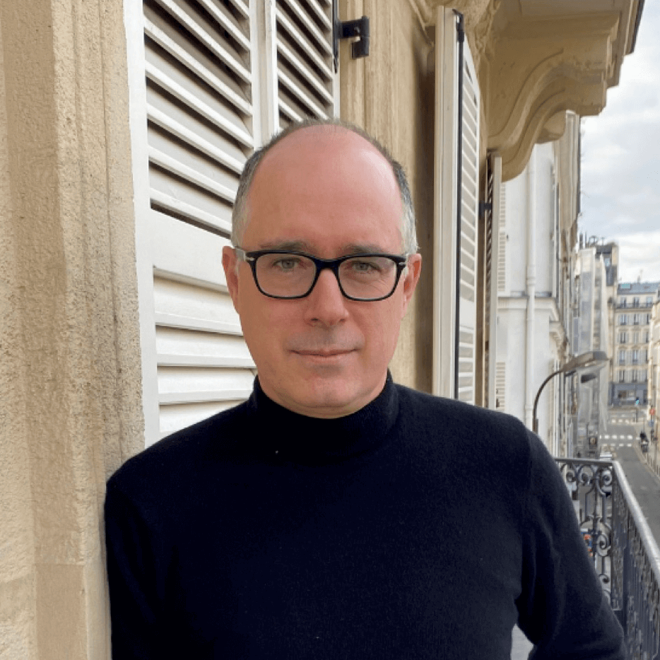 Un homme portant des lunettes et un col roulé noir se tient sur un balcon à côté de volets beiges, avec des immeubles et une rue visibles en arrière-plan.