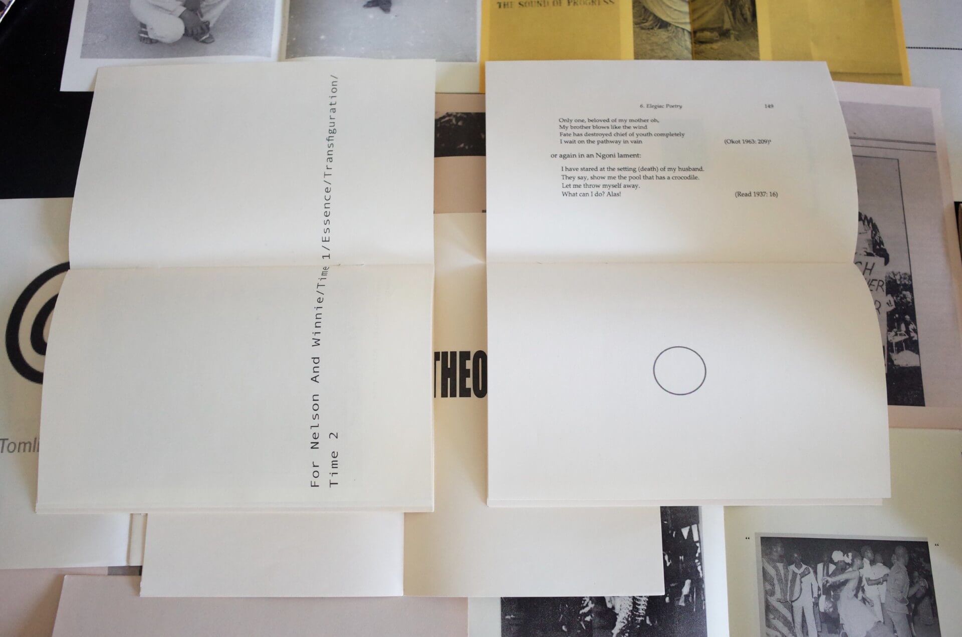 Deux livres ouverts sont posés sur une table avec divers papiers et livres en dessous. La page de gauche présente un texte aligné verticalement ; le livre de droite présente de la poésie et un petit cercle.