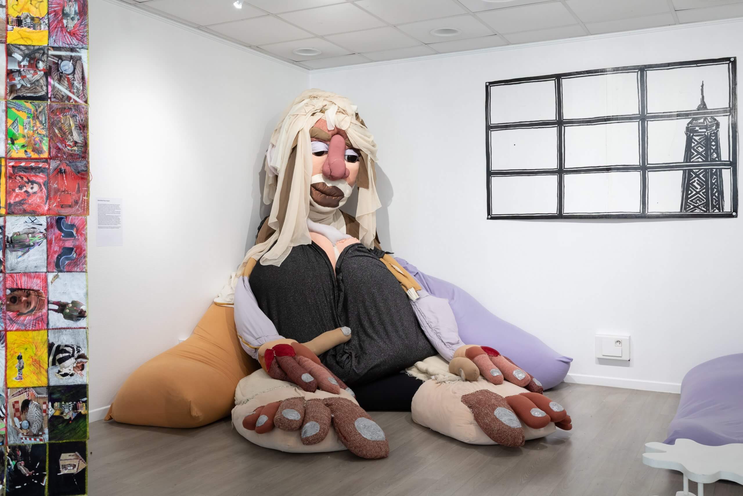 Une grande marionnette en tissu assise, au visage détaillé et aux mains et pieds surdimensionnés, est exposée dans un coin d'une salle.