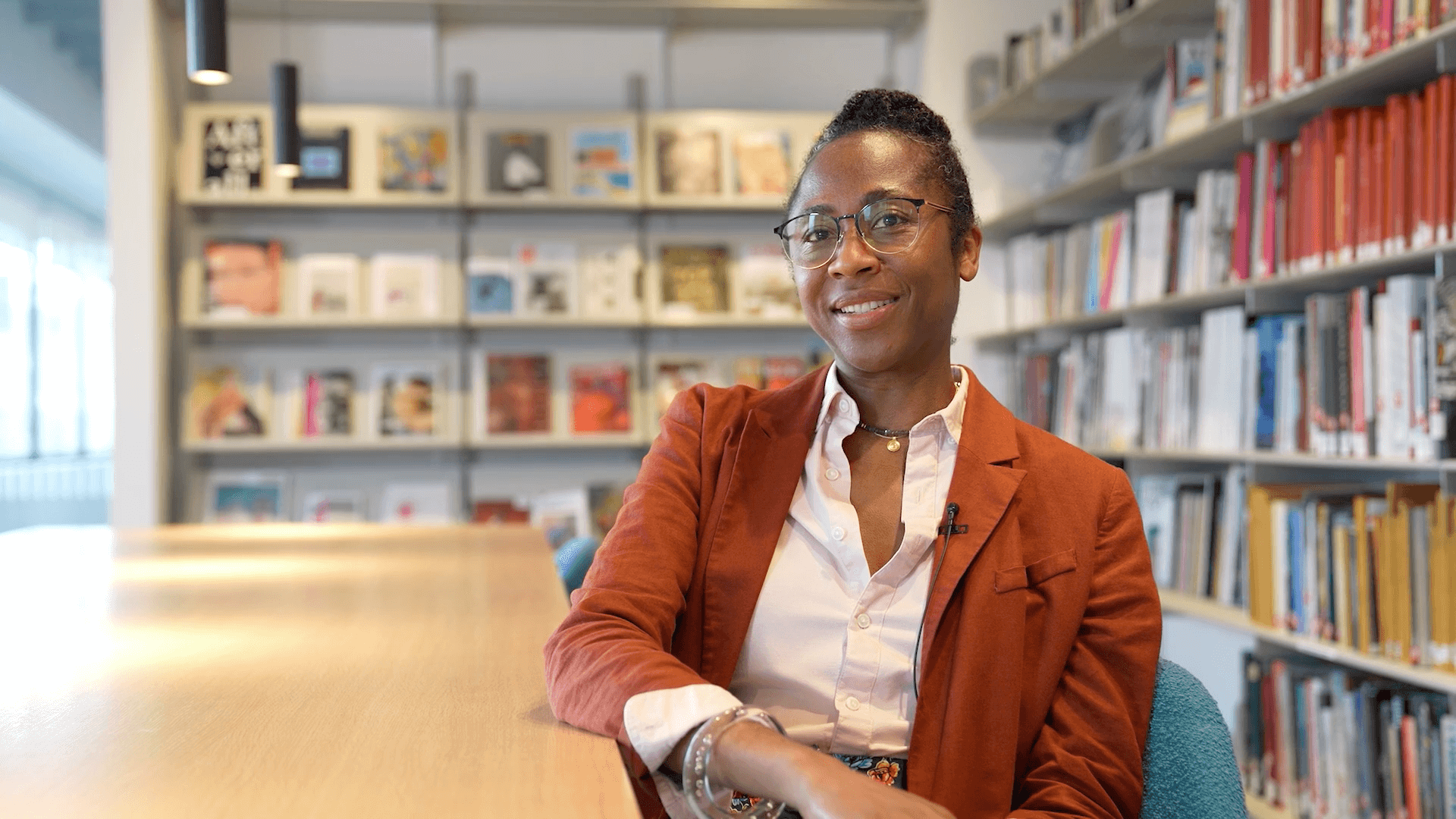 Naomi Beckwith est assise à une table dans une bibliothèque, avec des étagères de livres et de magazines en arrière-plan.