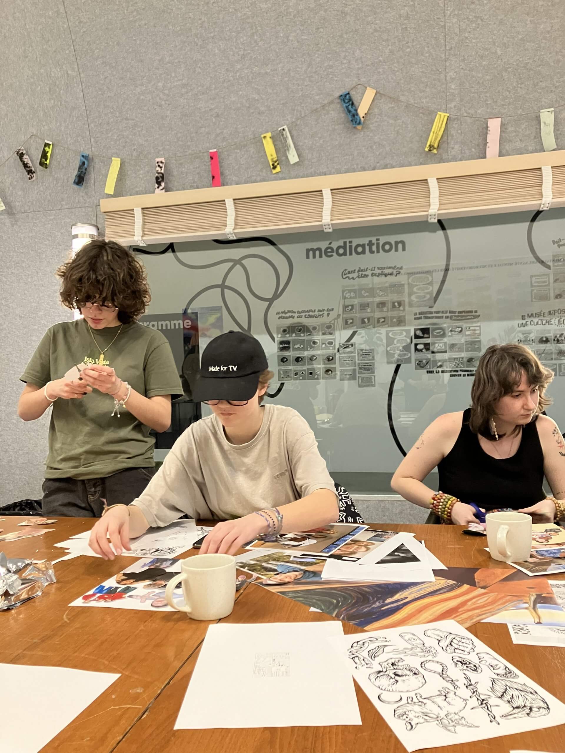 Trois personnes sont assises à une table et travaillent sur des projets artistiques avec des magazines, du papier et des tasses. Derrière eux se trouve une paroi vitrée où sont exposés divers papiers et dessins.