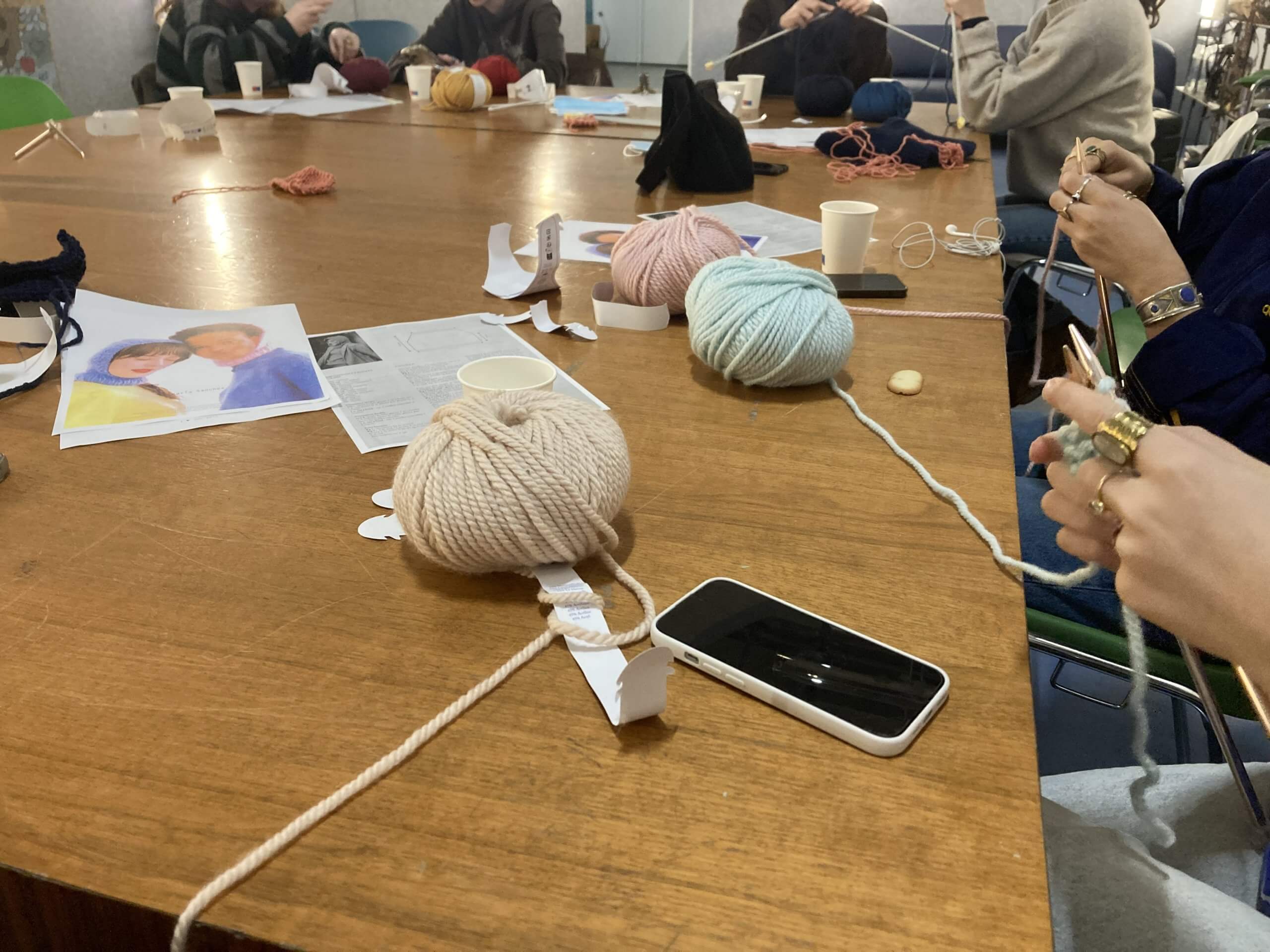Des personnes sont assises autour d'une table en bois avec du fil, des aiguilles à tricoter, un téléphone, des tasses et des documents imprimés, et participent à une activité de tricotage.