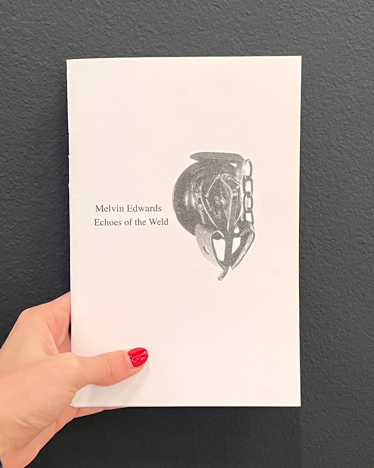 A hand with red nail polish holds a white booklet titled Melvin Edwards Echoes of the Weld featuring an abstract black and white artwork on the cover.