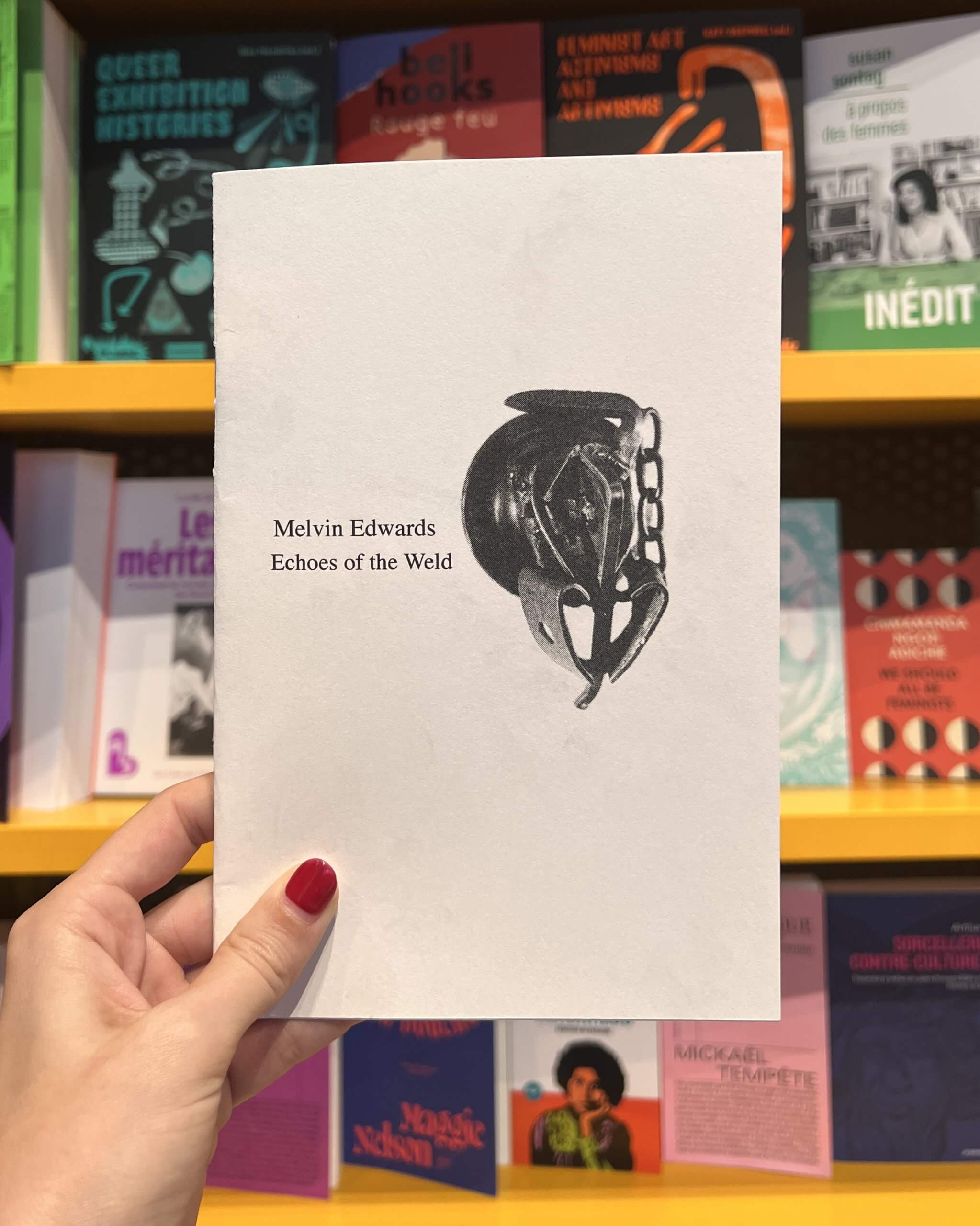 A hand with red nail polish holds a book titled Melvin Edwards Echoes of the Weld in front of a shelf filled with colorful books.