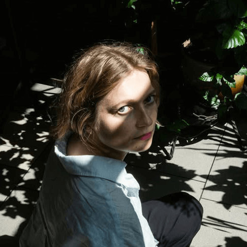 Une femme aux cheveux châtain clair est assise sur le sol dans une lumière solaire partielle, avec des ombres de feuilles sur son visage et un arrière-plan de plantes vertes.