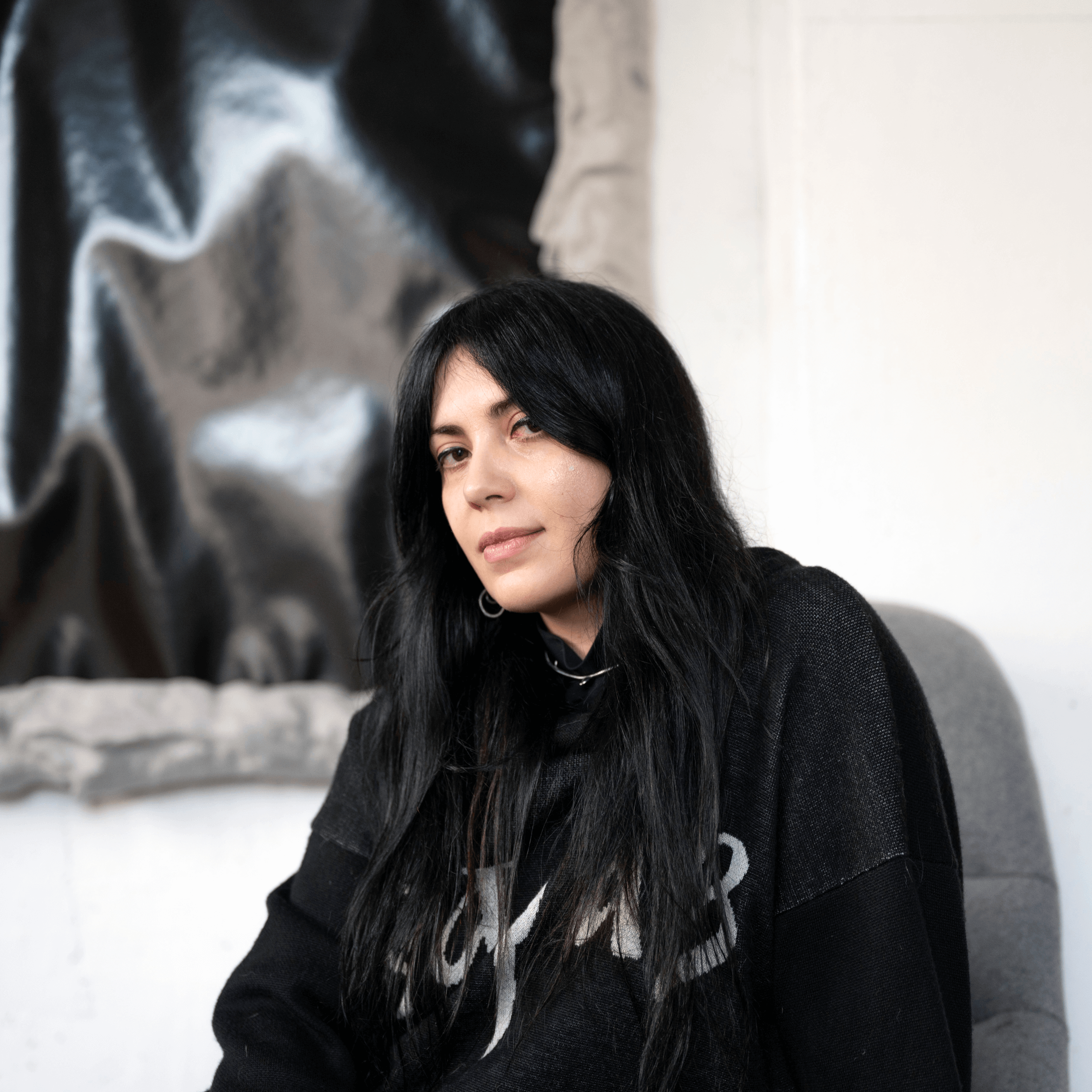 Une personne aux longs cheveux noirs est assise sur une chaise à l'intérieur, vêtue d'un pull noir. Une œuvre d'art abstraite noire et grise est accrochée au mur à l'arrière-plan.
