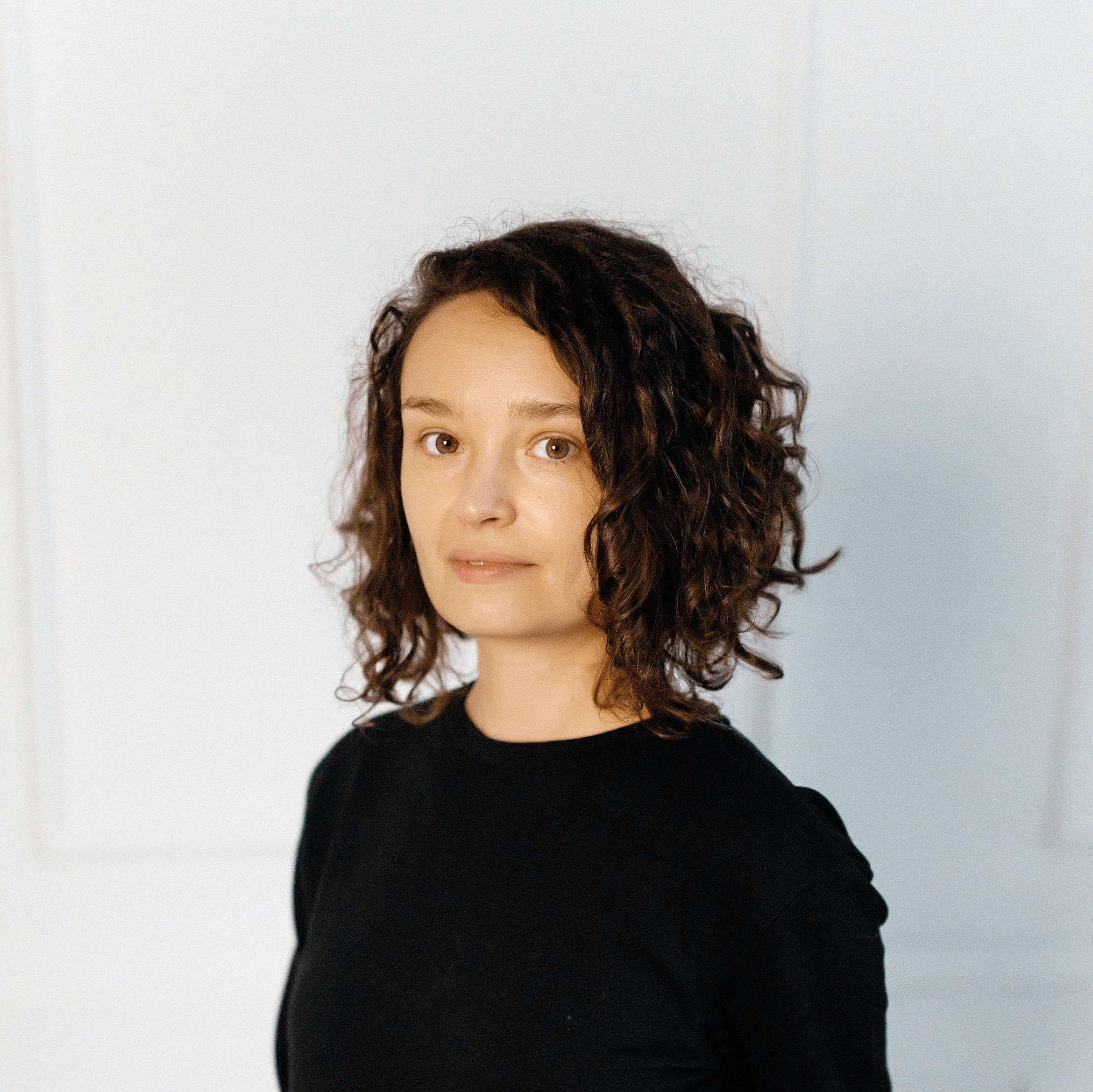 Une femme aux cheveux bruns bouclés, vêtue d'un haut noir, se tient devant un fond clair uni et regarde l'appareil photo avec une expression neutre.