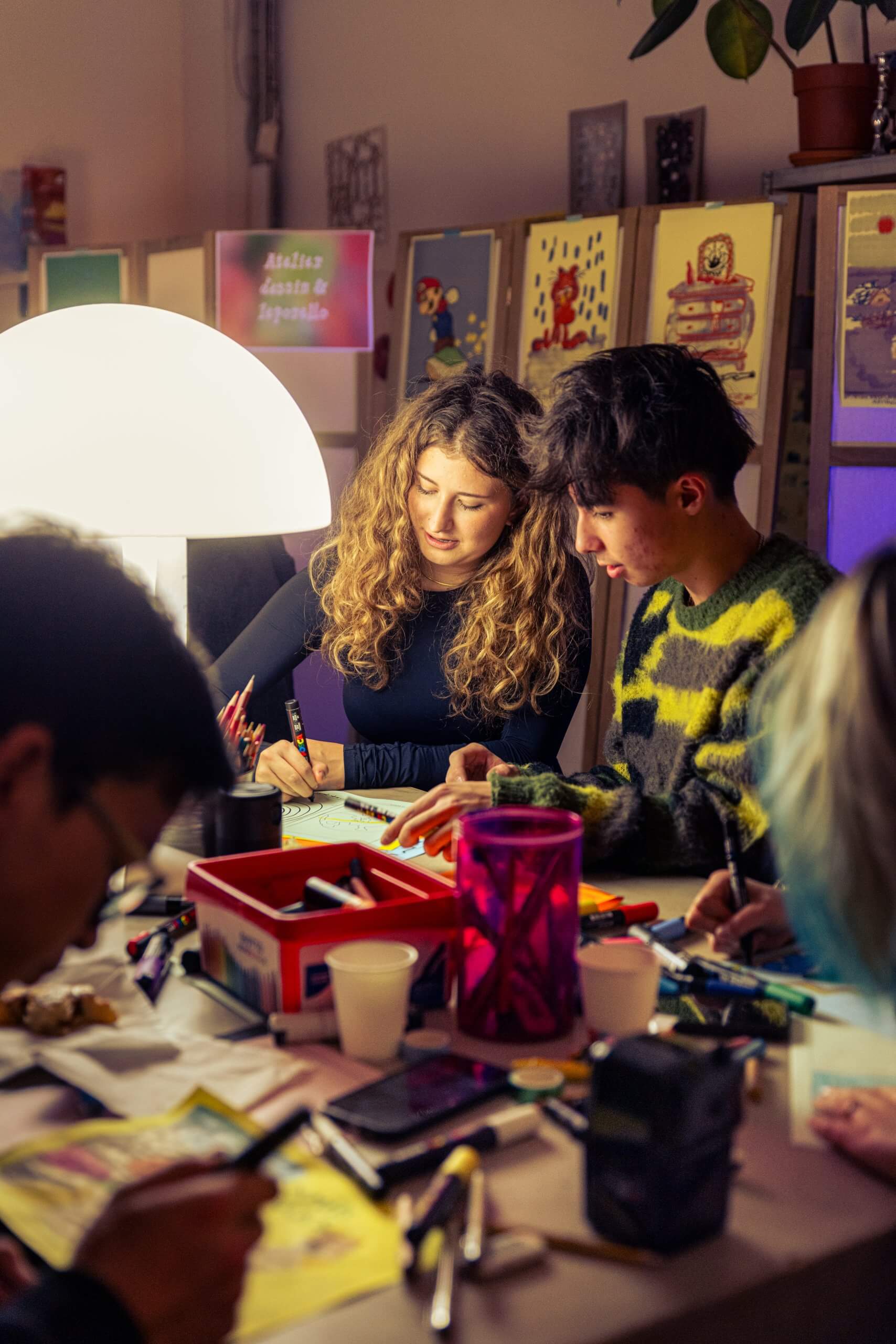 Plusieurs personnes sont assises autour d'une table couverte de fournitures artistiques, dessinant et travaillant sur des projets créatifs dans un espace intérieur bien éclairé.