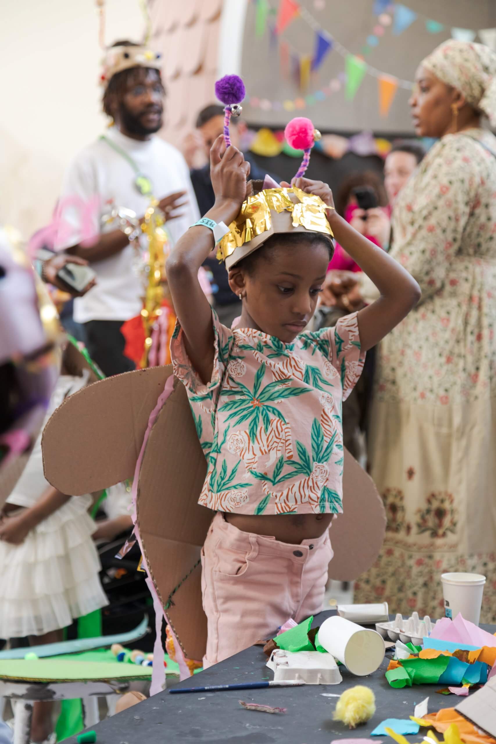 Un enfant portant une couronne en papier avec des antennes et des ailes en carton ajuste son costume à une table de bricolage lors d'un événement festif en intérieur.