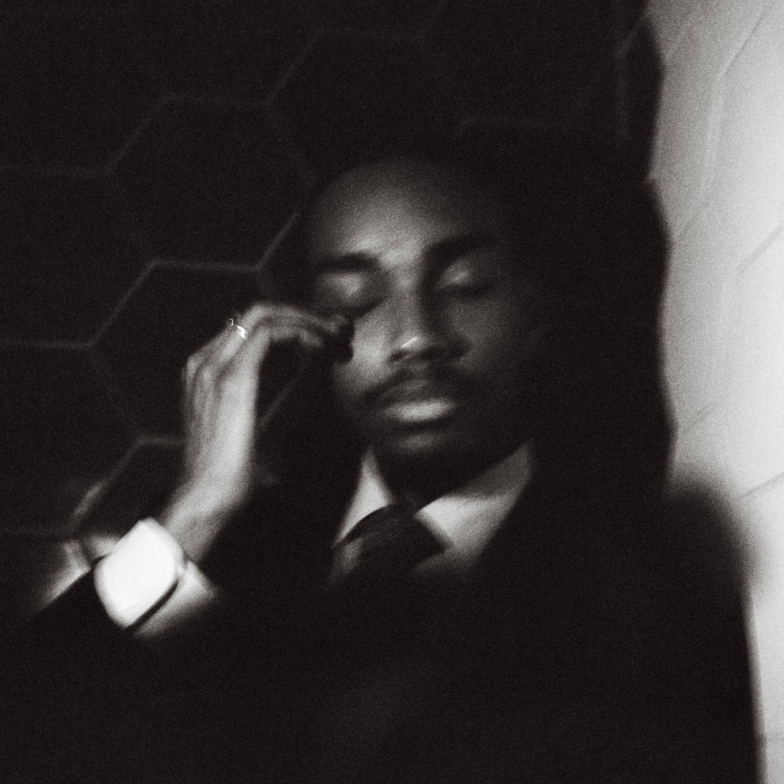 Un homme en costume est assis, les yeux fermés, se touchant le visage, dans un environnement faiblement éclairé, avec un mur à motifs hexagonaux en arrière-plan. L'image est en noir et blanc et légèrement floue.