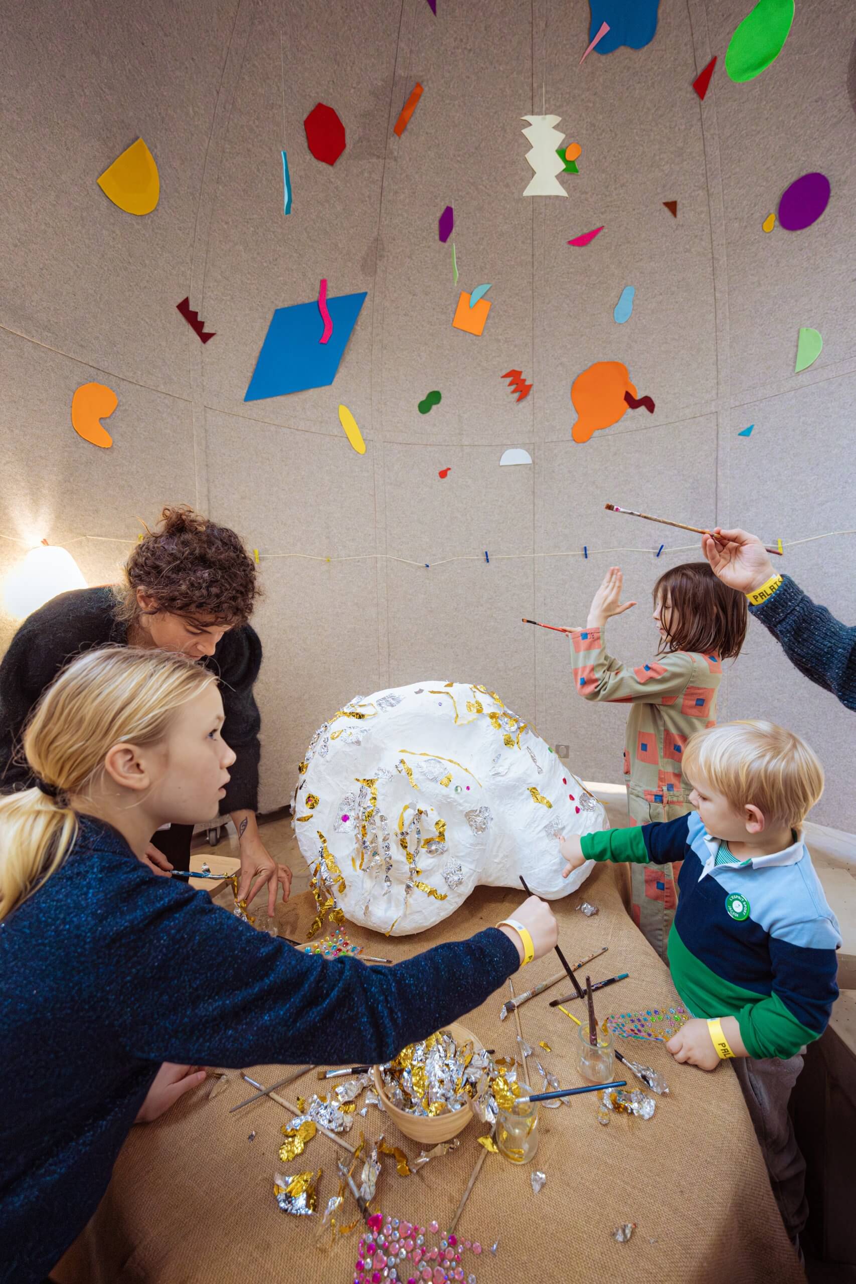 Des enfants et des adultes décorent une grande sculpture en papier blanc avec de la peinture et des paillettes dans une pièce où des formes colorées sont suspendues au plafond.