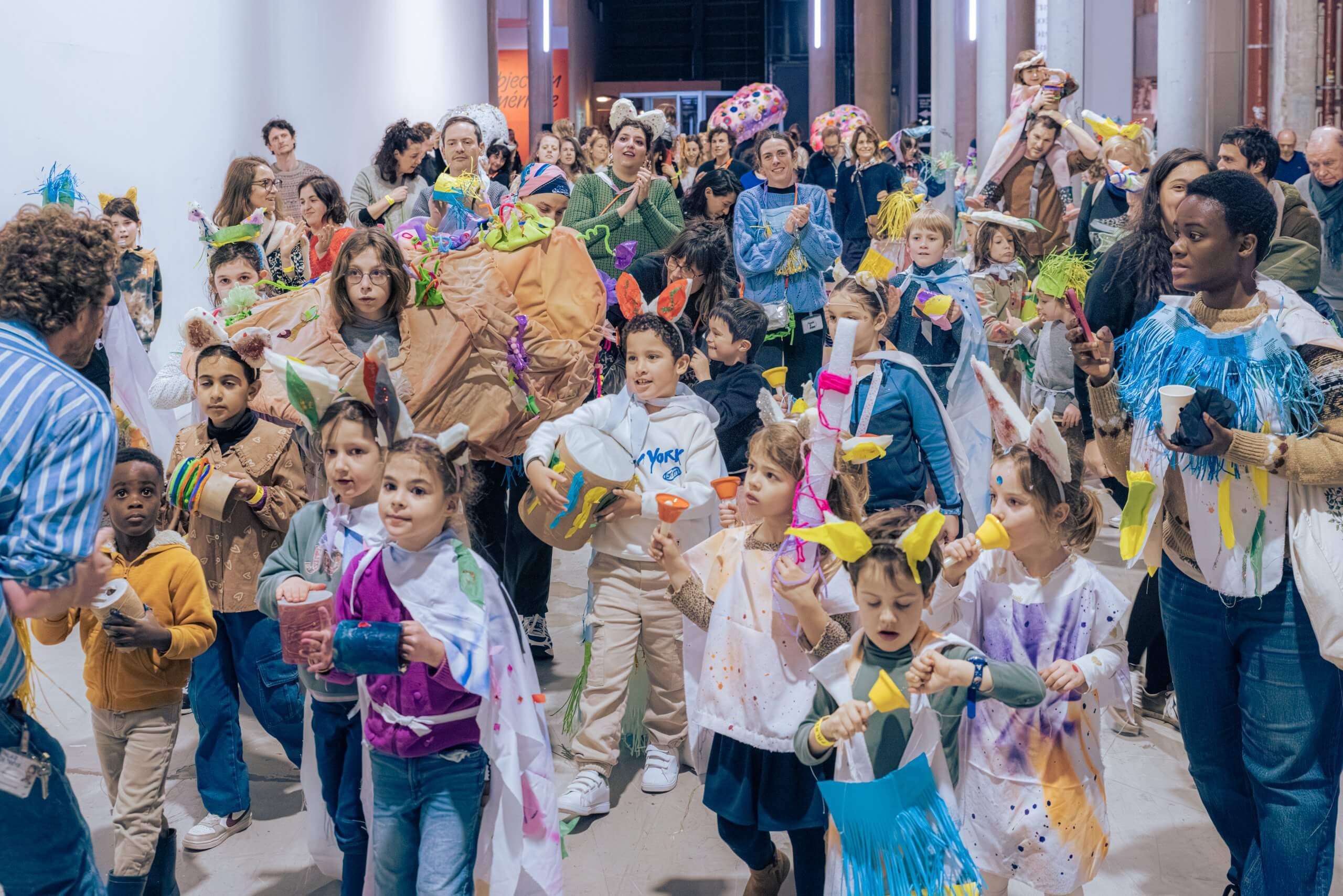 Un grand groupe d'enfants et d'adultes, dont certains portent des costumes colorés, se rassemblent à l'intérieur pour ce qui semble être un événement festif ou communautaire.