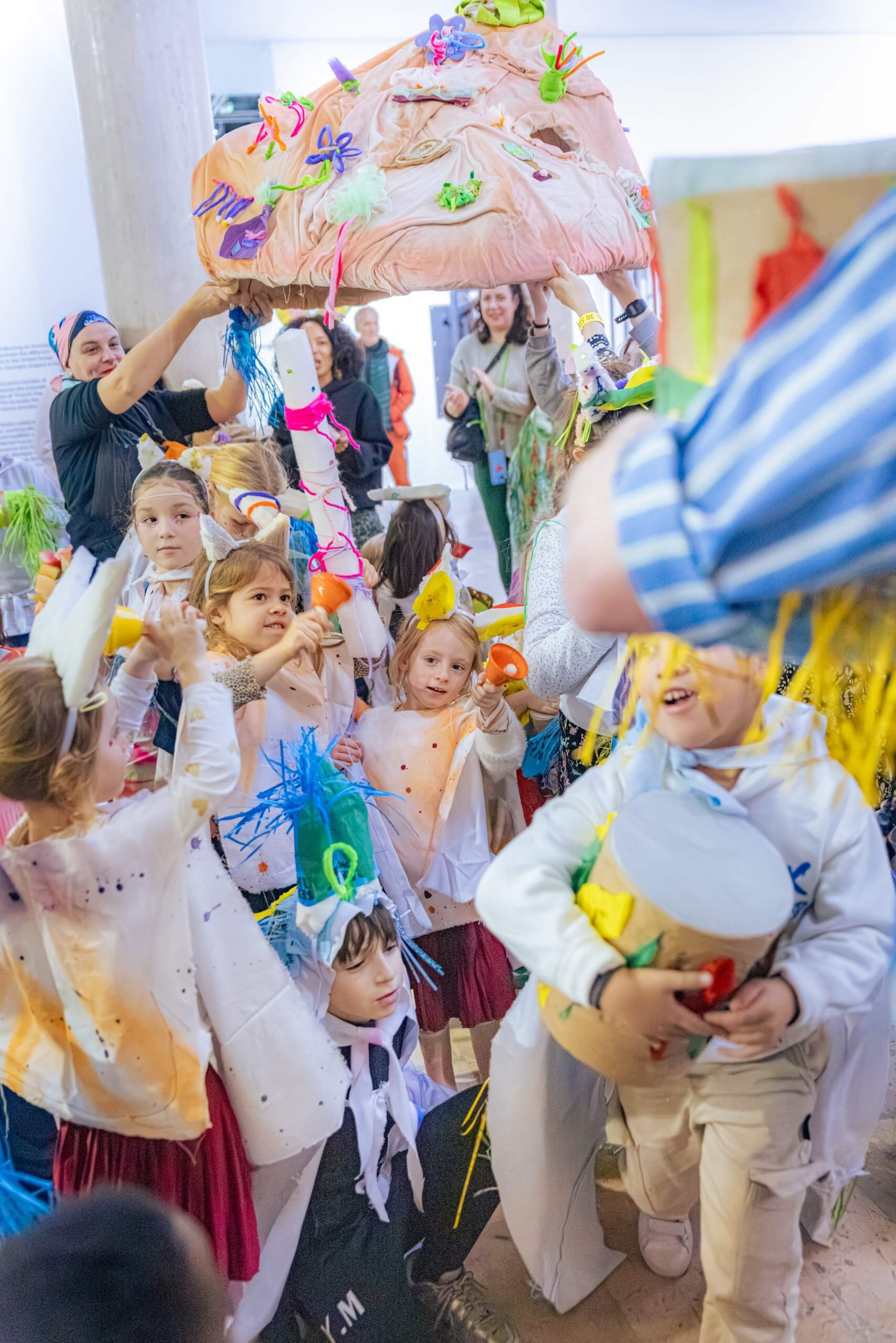 Un groupe d'enfants vêtus de costumes colorés faits à la main participe à un événement en intérieur, avec l'aide d'adultes et des décorations au-dessus de leur tête.