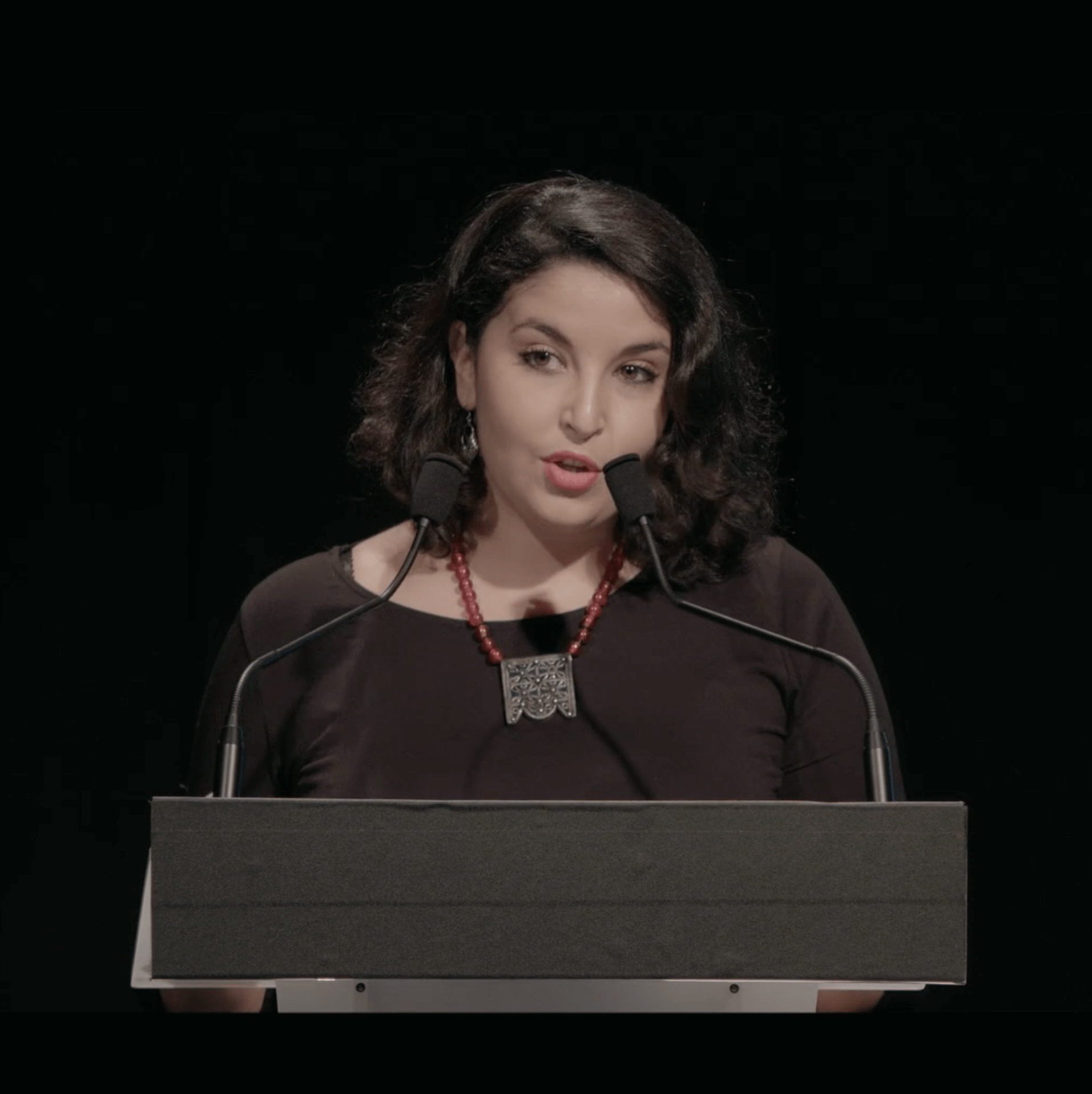 Une femme aux cheveux noirs s'exprime sur un podium muni de deux microphones. Elle porte un haut noir, un collier de perles rouges et un grand pendentif, sur un fond sombre.
